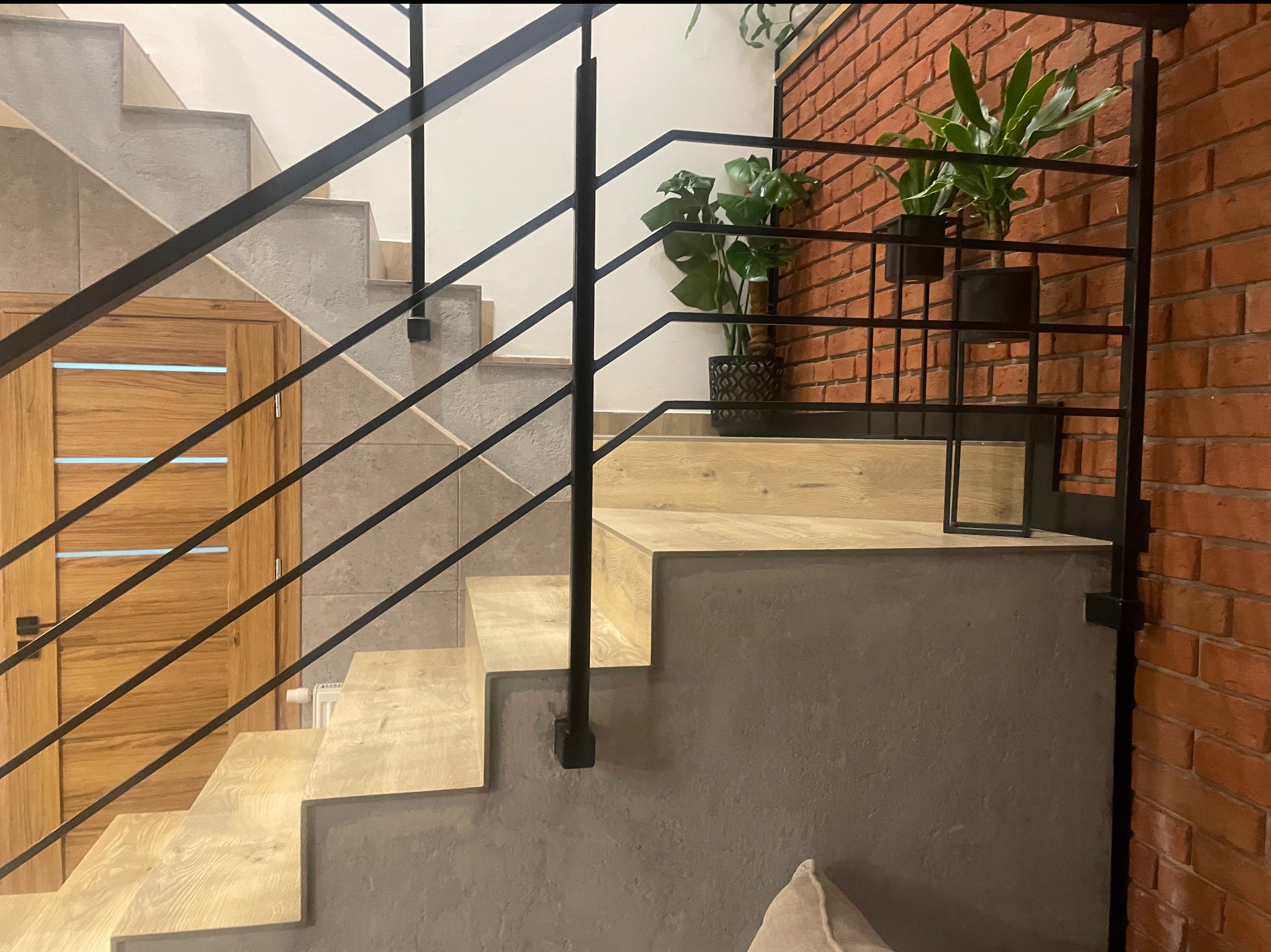 Nowoczesne schody z drewnianymi stopniami i czarną, minimalistyczną balustradą z metalowych prętów, na tle ceglanej ściany z roślinami doniczkowymi.