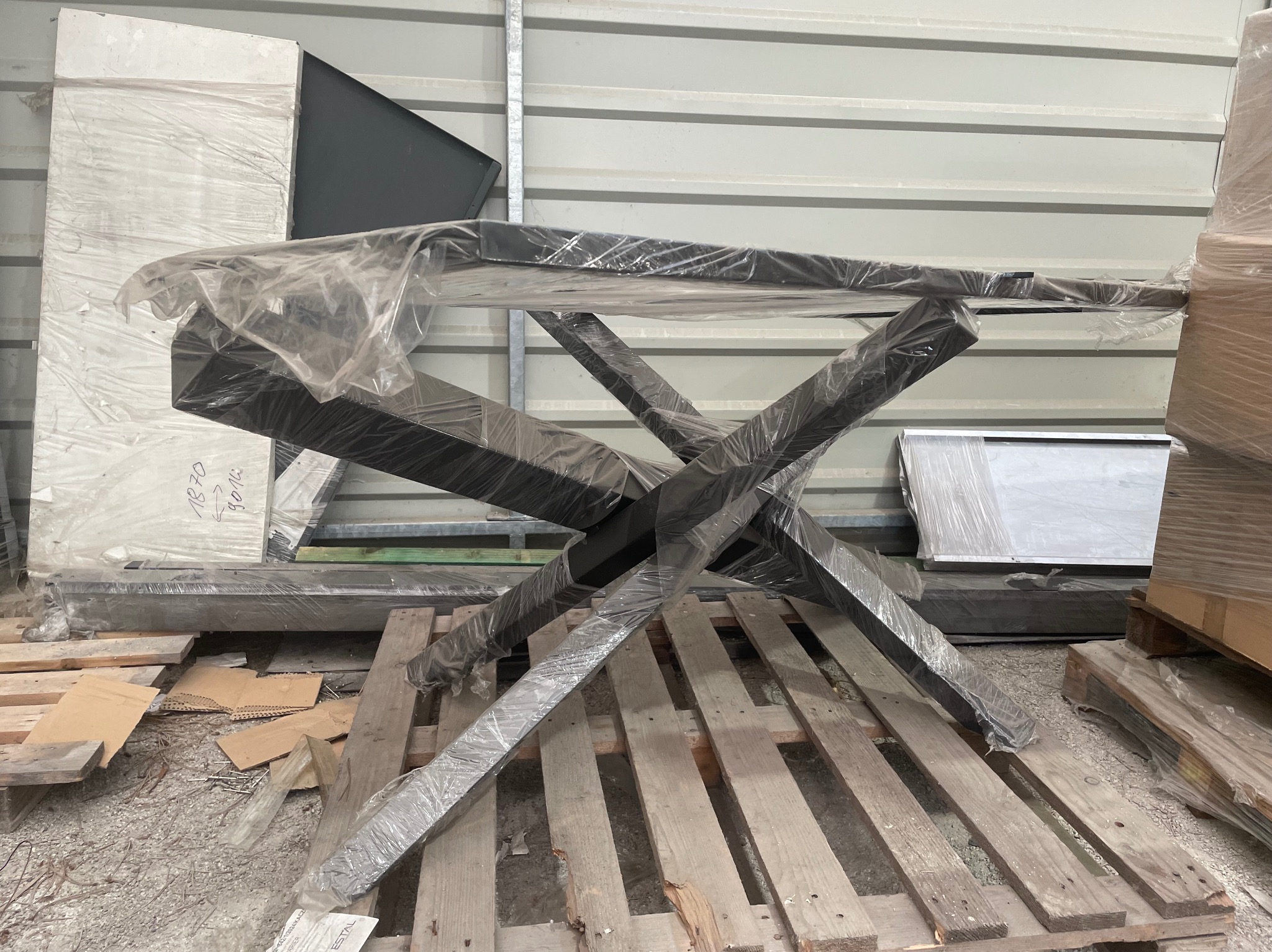 Metalowa konstrukcja stołu z krzyżowymi nogami, zabezpieczona folią, leży na palecie w magazynie. Elementy metalowe w kolorze czarnym.