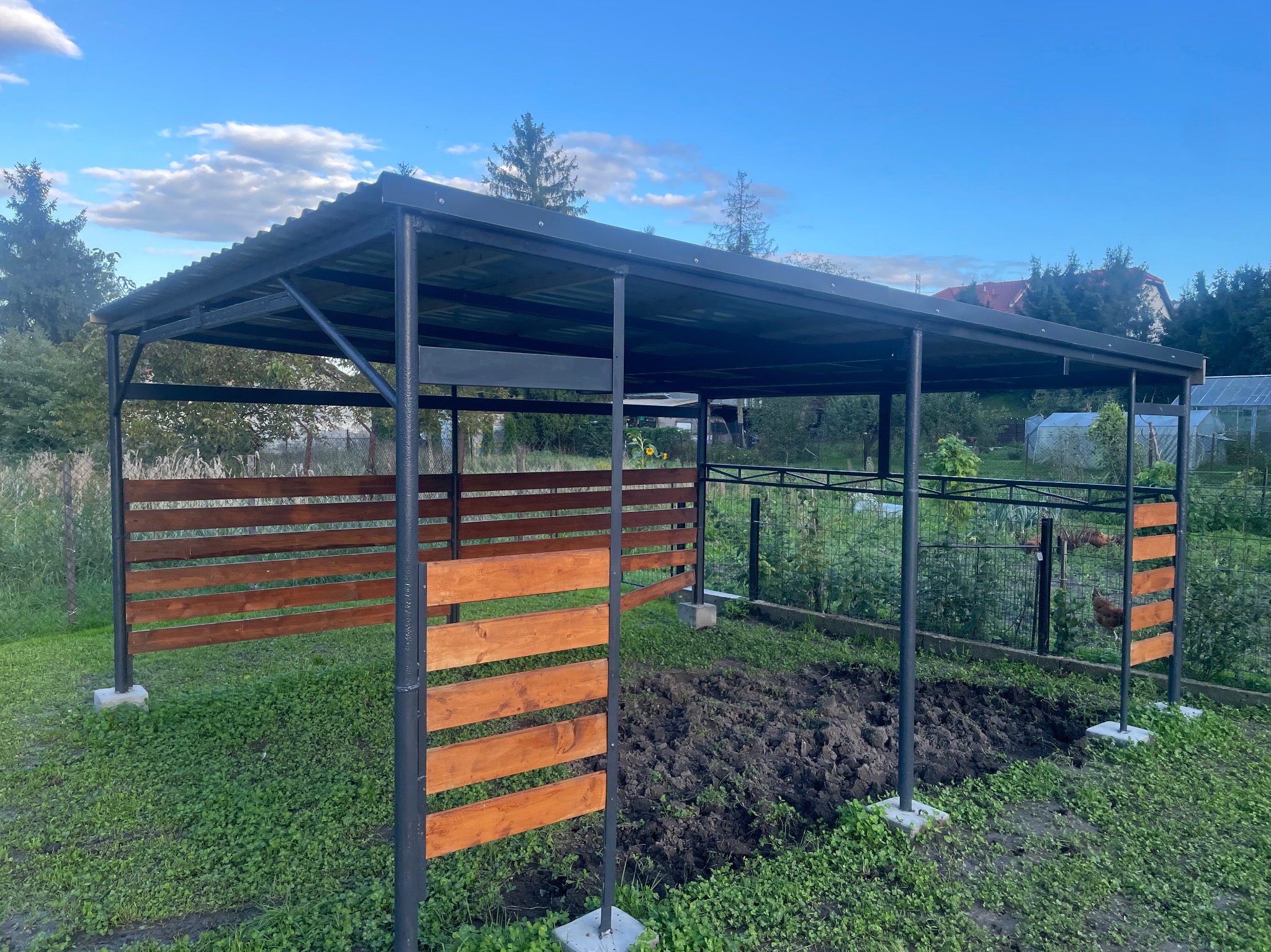 Metalowa konstrukcja z drewnianymi panelami w ogrodzie, prawdopodobnie wiata lub pergola. Konstrukcja pomalowana na czarno, panele drewniane w ciepłym odcieniu brązu.