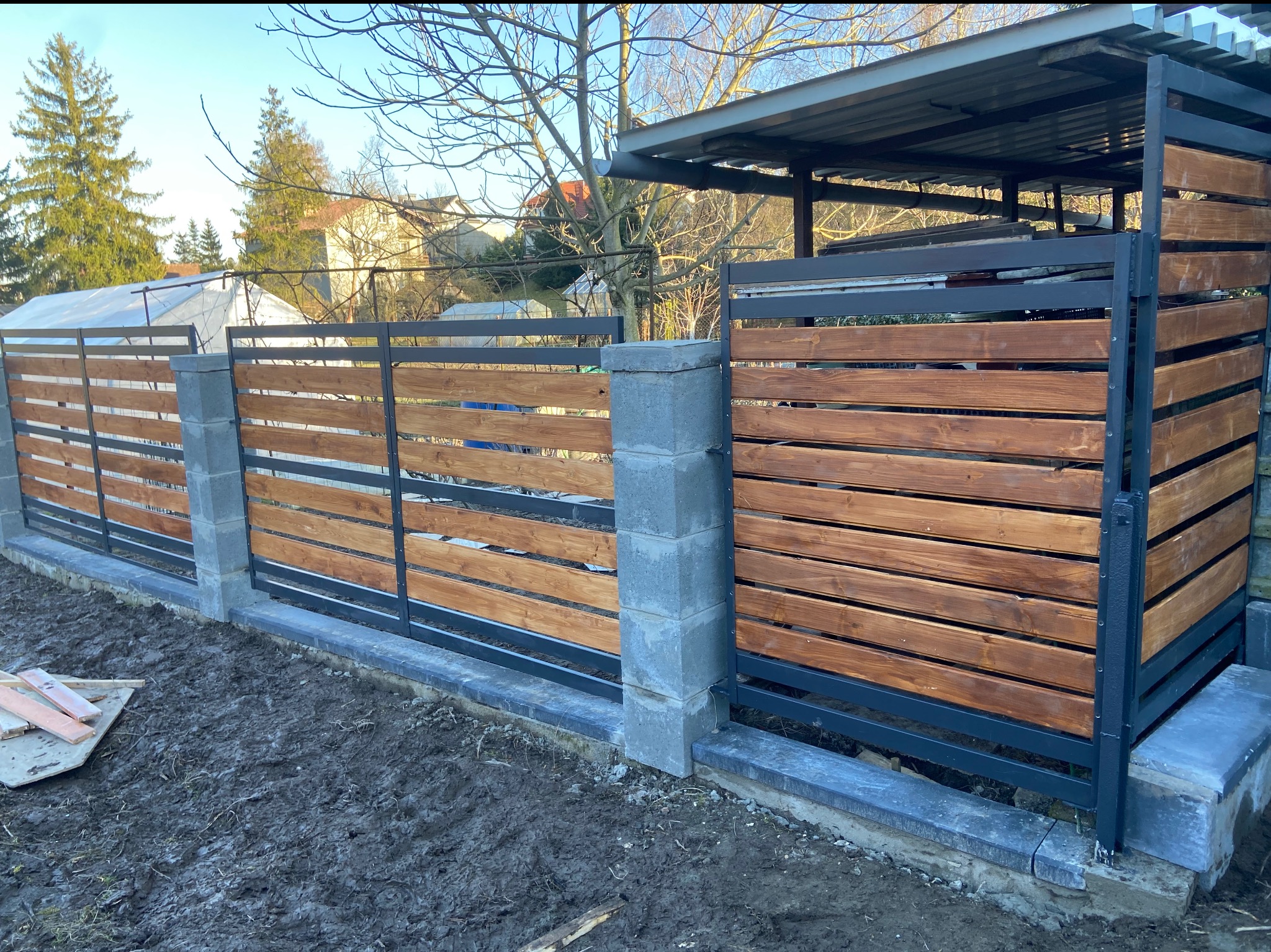 Nowoczesne ogrodzenie z drewnianymi panelami i metalową ramą, osadzone na betonowych słupkach. Widoczne elementy konstrukcyjne i surowe wykończenie terenu.