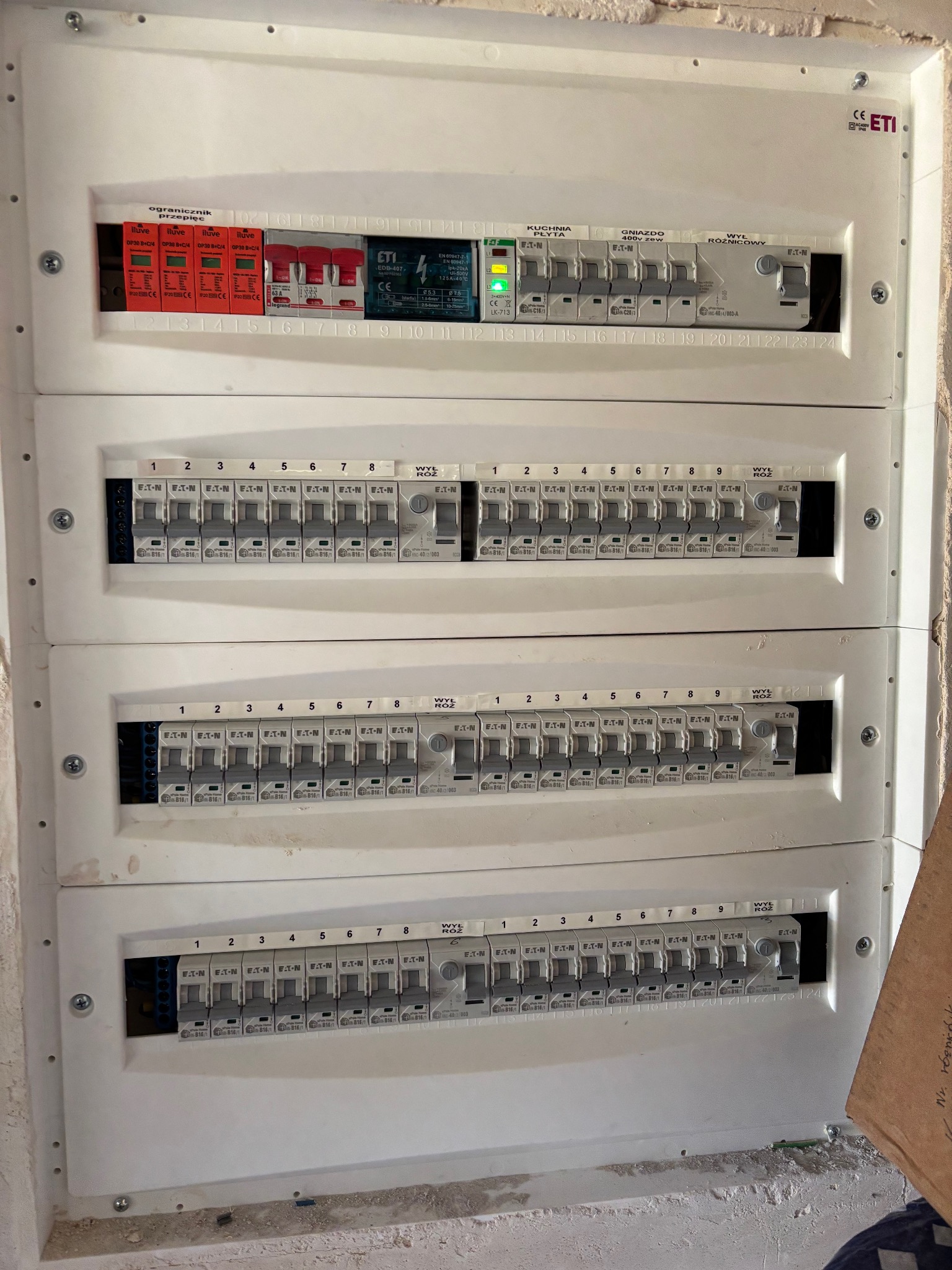 Rozdzielnica elektryczna z zabezpieczeniami i wyłącznikami różnicowoprądowymi, zamontowana na ścianie. Widoczne oznaczenia i numery obwodów.