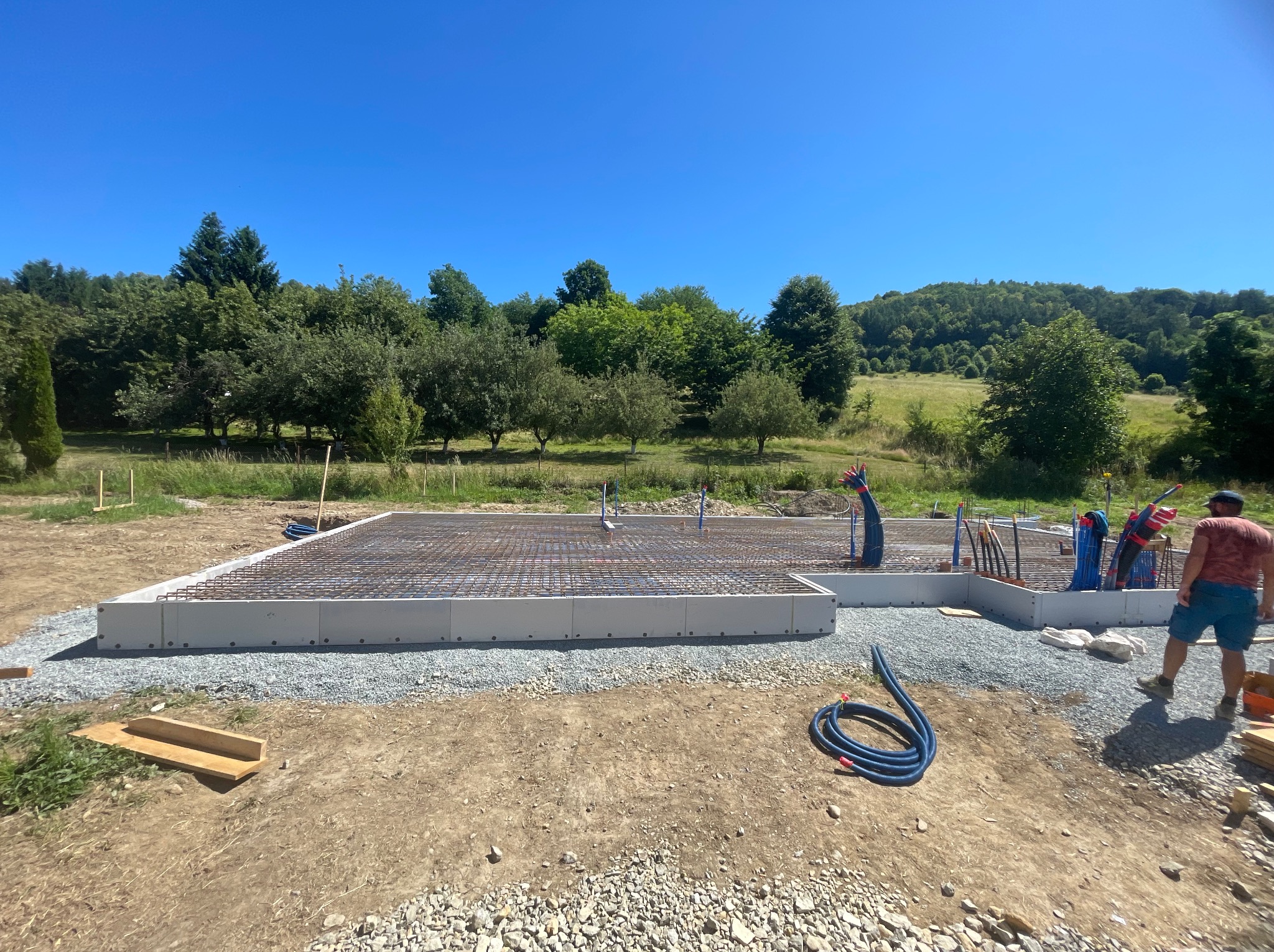 Zbrojona płyta fundamentowa na żwirowym podłożu, widoczne instalacje, w tle zieleń i błękitne niebo. Fundament pod budowę domu jednorodzinnego w słoneczny dzień.