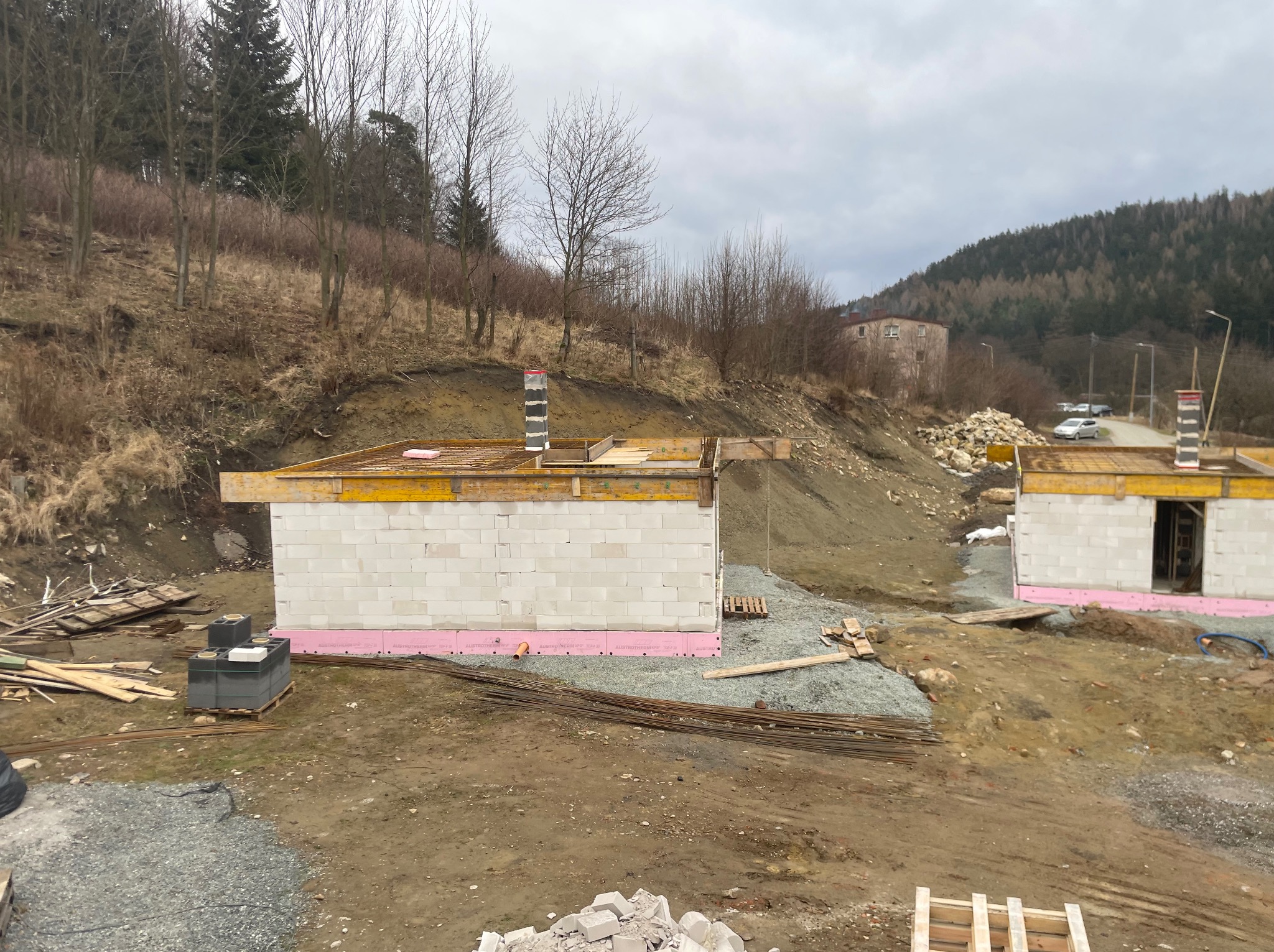 Budowa domu w toku: ściany z białych bloczków, różowa izolacja fundamentów, drewniane szalunki, zbrojenie. W tle wzgórze z drzewami i budynek.