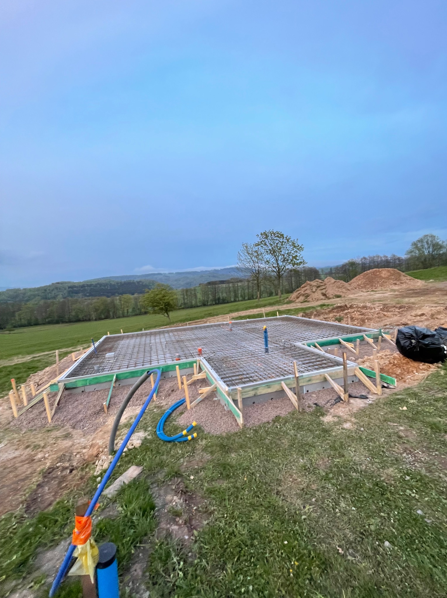 Zbrojona płyta fundamentowa z szalunkiem na działce z widokiem na wzgórza, rury instalacyjne w ziemi, pochmurne niebo. Etap budowy domu.