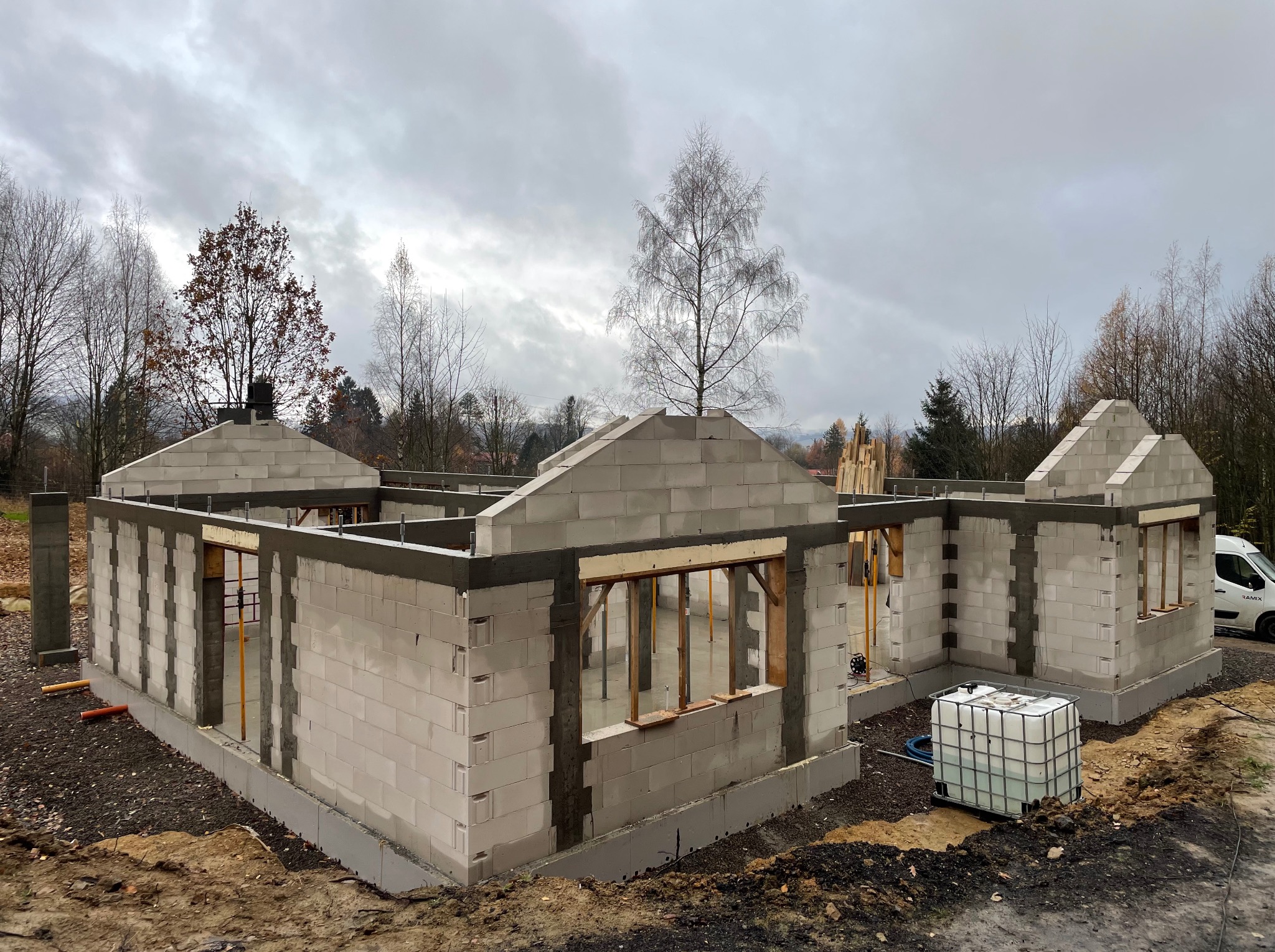 Surowy stan domu z betonu komórkowego w trakcie budowy. Widoczne ściany, otwory okienne z drewnianymi stemplami i fundamenty. W tle drzewa i pochmurne niebo.