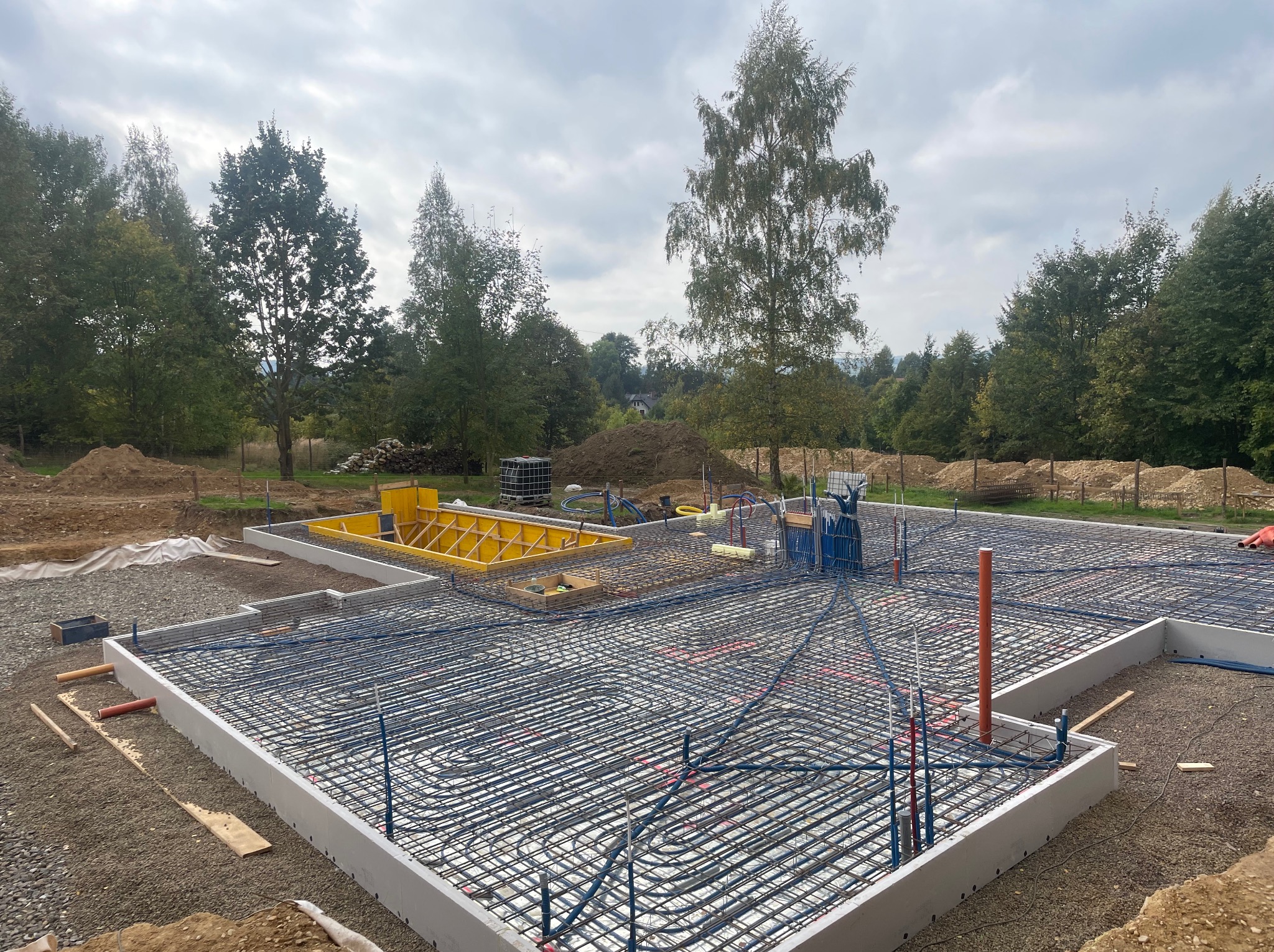 Zbrojenie fundamentu z instalacją ogrzewania podłogowego wewnątrz szalunku, widok z góry na plac budowy z drzewami w tle, pochmurne niebo.