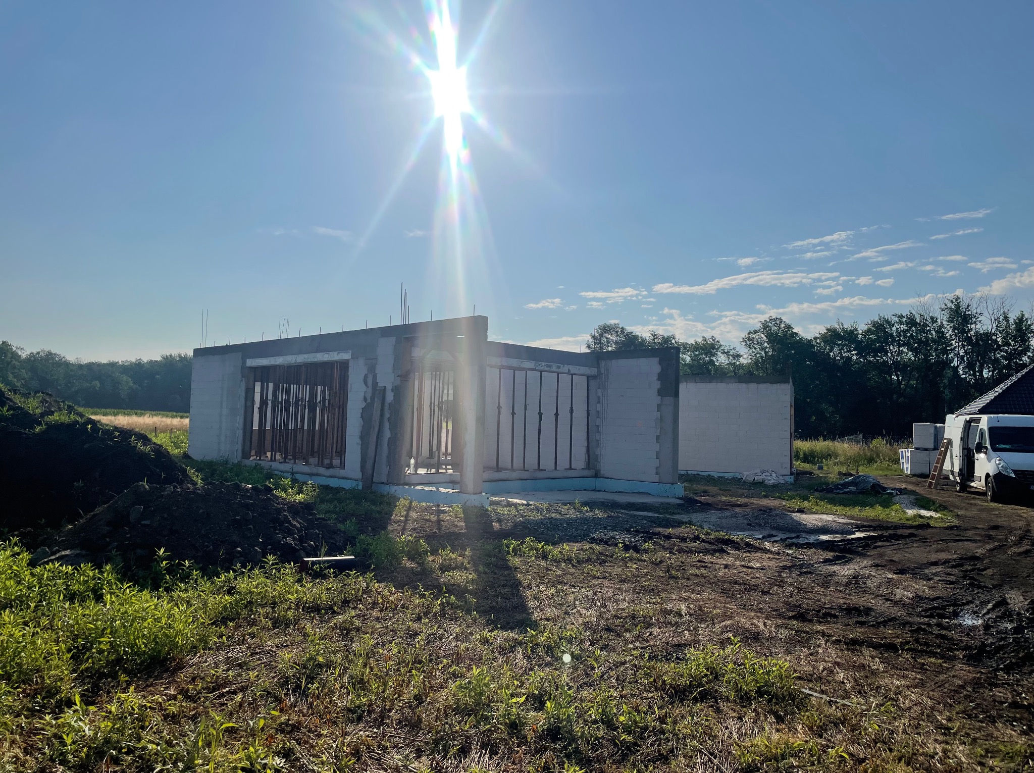 Wczesna faza budowy domu z betonu komórkowego w słoneczny dzień. Widoczne ściany, stalowe elementy konstrukcyjne, ziemia i pojazd dostawczy w tle.