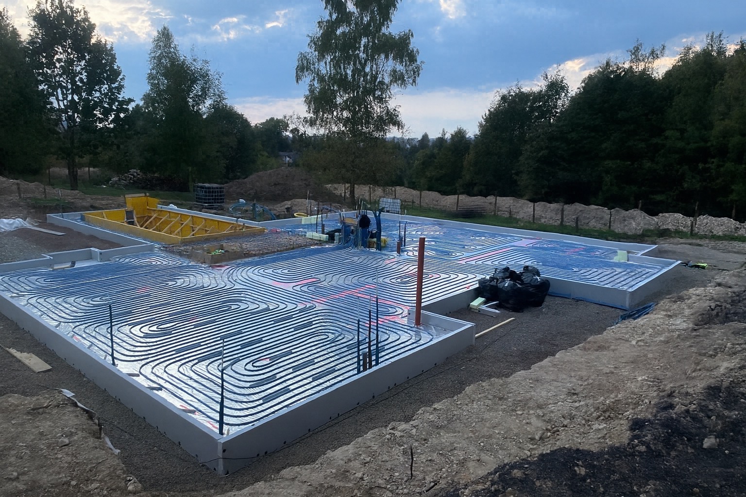 Fundamenty domu z ułożonym systemem ogrzewania podłogowego. Widoczne niebieskie rury w kształcie meandrów na izolacji. W tle drzewa i niebo.