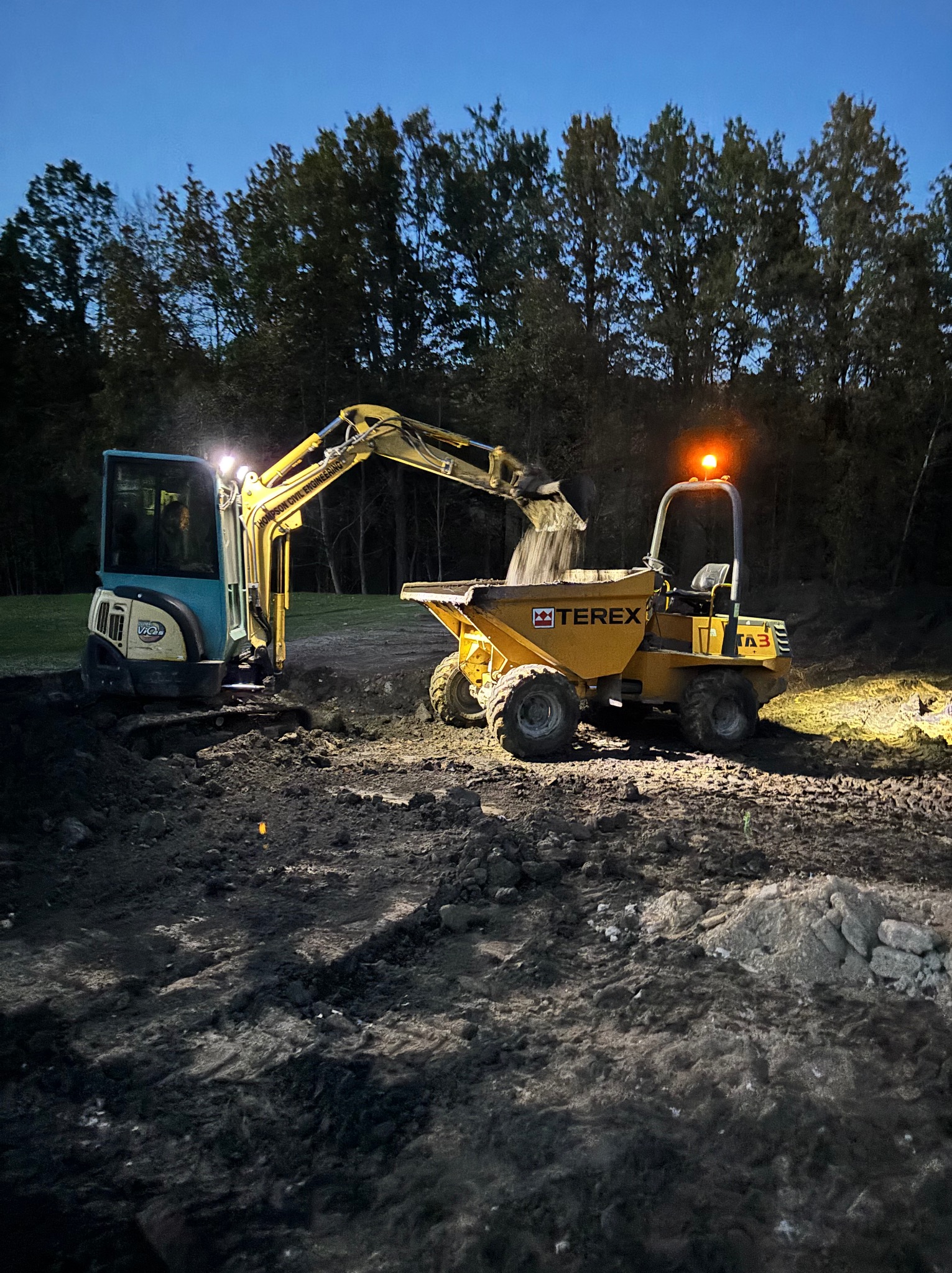 Nocne roboty ziemne: minikoparka i wywrotka Terex oświetlają teren podczas wykopu. Widoczne drzewa w tle i świeżo naruszona ziemia.