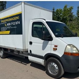 GRAND-Przeprowadzki - Biały samochód dostawczy Renault Master z reklamą firmy przeprowadzkowej, zaparkowany na ulicy w słoneczny dzień. Widoczny numer telefonu i adres strony internetowej.