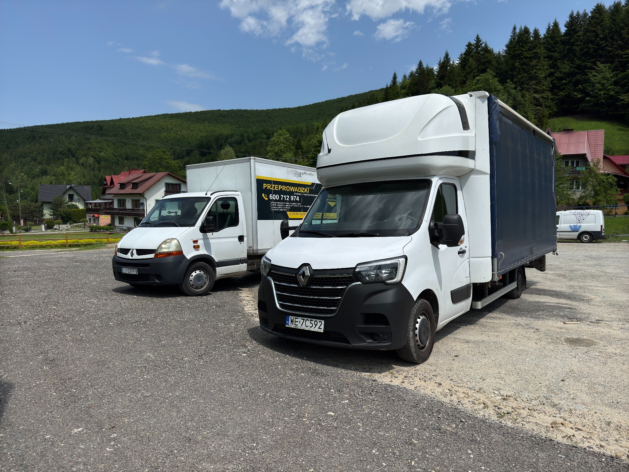 Dwa białe busy transportowe Renault na żwirowym parkingu z zielonym wzgórzem w tle. Jeden z busów ma plandekę i nadbudowę kabiny.