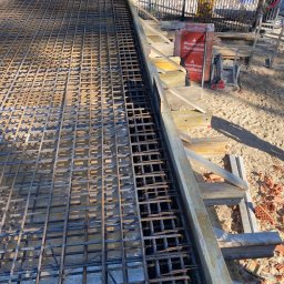 WAL-BUD - Siatka zbrojeniowa w szalunku fundamentu, widok z góry. Na drugim planie cegły Porotherm i drewniane elementy konstrukcyjne. Słoneczny dzień.