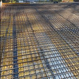 WAL-BUD - Siatka zbrojeniowa na żółtym szalunku, przygotowana do zalania betonem. Widok z góry, w tle budynek mieszkalny i zieleń.