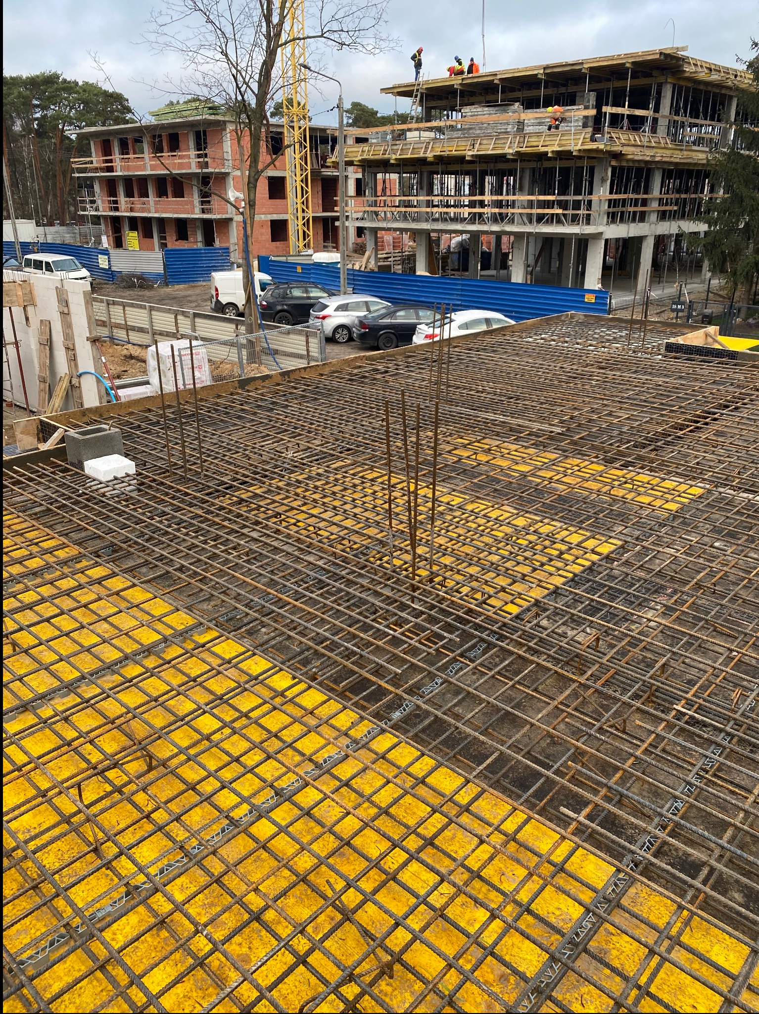 Siatka zbrojeniowa na żółtym szalunku, przygotowana do zalania betonem. W tle budynek w trakcie budowy, widoczny żuraw i pracownicy na górze konstrukcji.