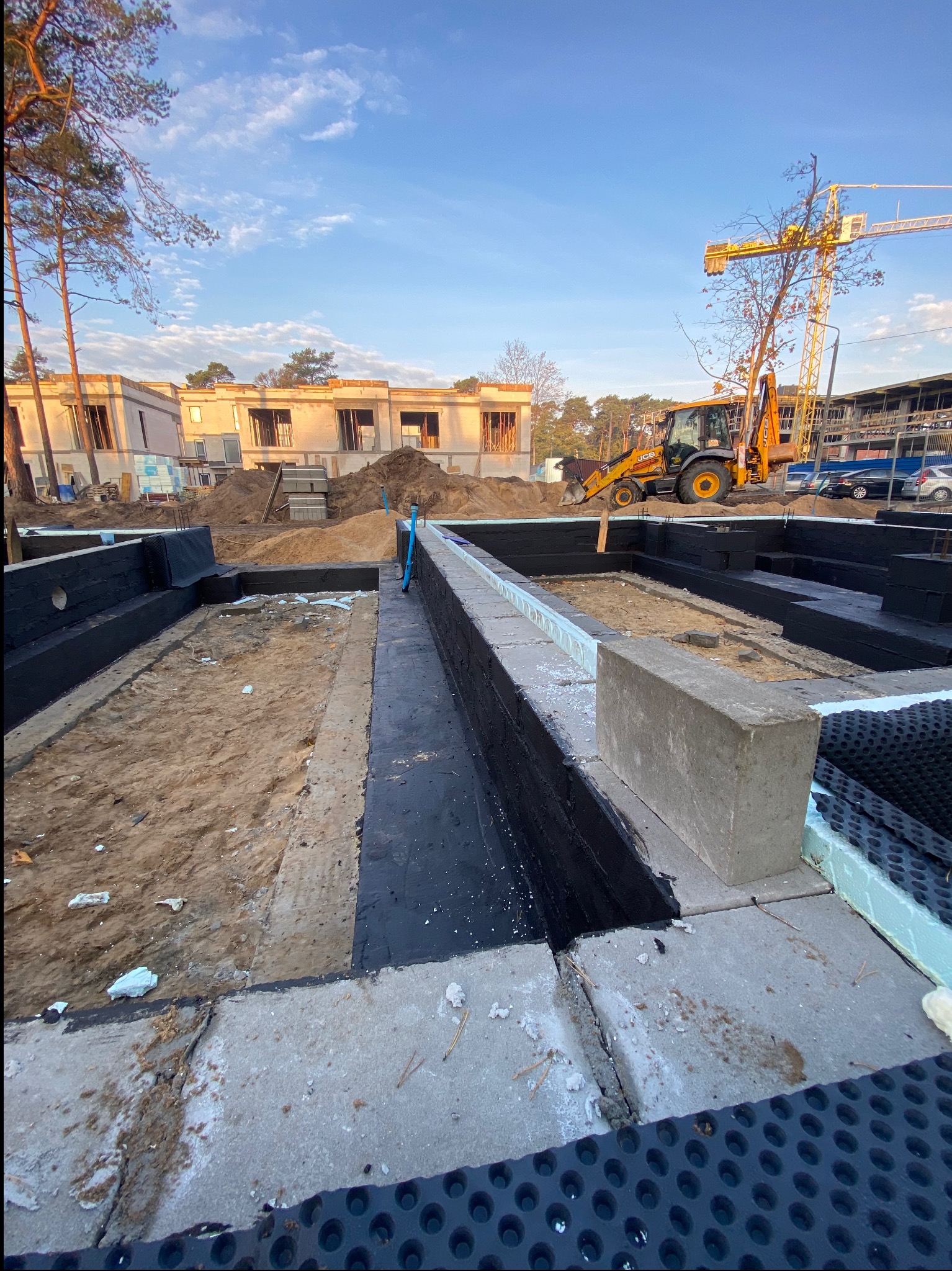 Fundamenty z izolacją pionową i drenażem w wykopie, w tle budynek w stanie surowym otwartym i koparko-ładowarka JCB. Widok na plac budowy w słoneczny dzień.
