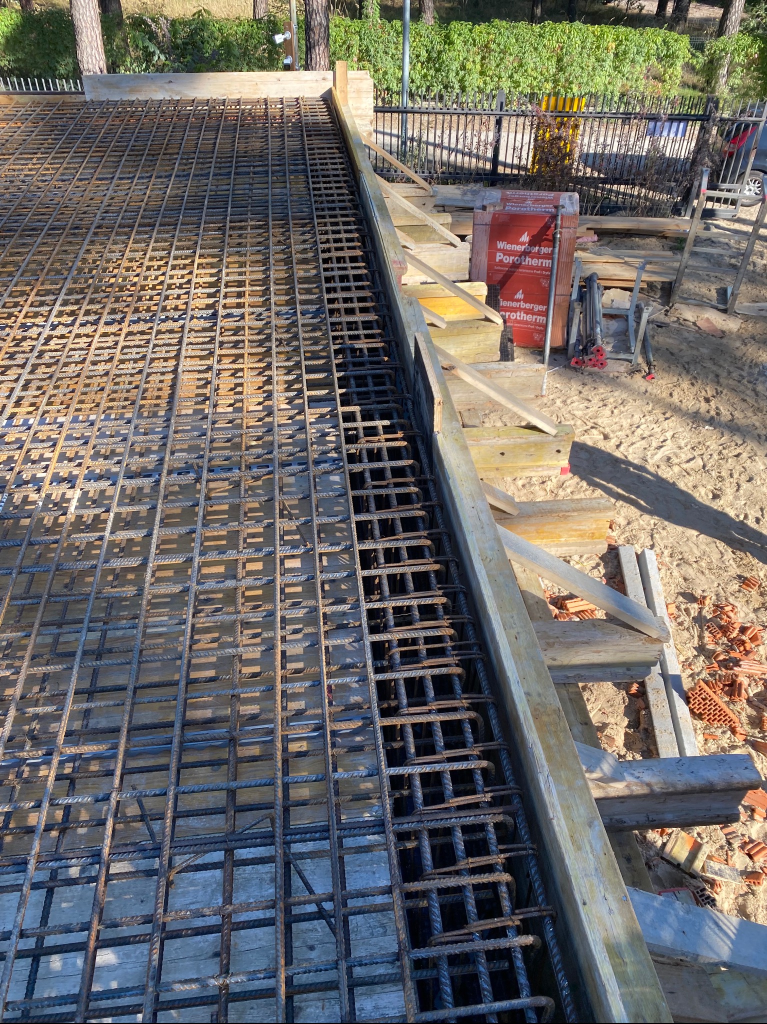 Siatka zbrojeniowa w szalunku fundamentu, widok z góry. Na drugim planie cegły Porotherm i drewniane elementy konstrukcyjne. Słoneczny dzień.