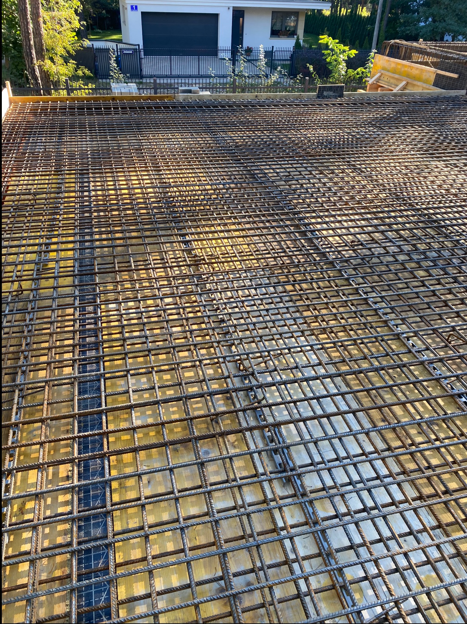 Siatka zbrojeniowa na żółtym szalunku, przygotowana do zalania betonem. Widok z góry, w tle budynek mieszkalny i zieleń.