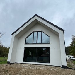 ArtBud - Nowoczesny dom z białymi ścianami i czarnymi ramami okiennymi. Charakterystyczne trójkątne okno na szczycie. Elewacja świeżo pomalowana, teren wokół w trakcie zagospodarowania.
