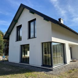 ArtBud - Nowoczesny, biały dom z dużymi oknami i czarnym dachem, otoczony zielenią i niebieskim niebem. Widoczne detale wykończenia elewacji i otoczenia.