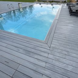 ArtBud - Nowoczesny taras z szarej deski kompozytowej otaczający basen z błękitną wodą. W tle widoczne drzewa i góry, tworzące malowniczy krajobraz.