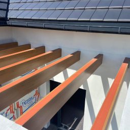 ArtBud - Drewniana pergola z pomarańczowymi akcentami, widok z dołu na tle białej ściany i fragmentu dachu z czarnej dachówki. Widoczne ocieplenie między belkami.