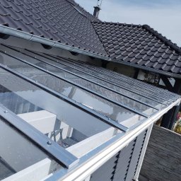 ArtBud - Szklana zabudowa tarasu z metalową konstrukcją, widok z góry, z fragmentem dachu domu w tle. Nowoczesne rozwiązanie zadaszenia.