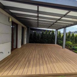 ArtBud - Nowoczesny taras z drewnianą podłogą i metalową pergolą, otoczony zielenią. Widok na dom z dużymi oknami i starannie utrzymany ogród z tujami.