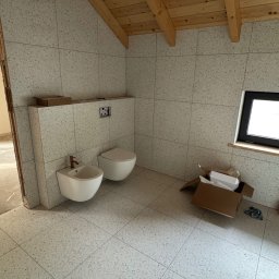 ArtBud - Wykończona łazienka na poddaszu z beżowymi płytkami, widoczne białe sanitariaty, drewniana więźba dachowa i okno. Częściowo widoczny karton po armaturze.