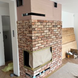 ArtBud - Wnętrze w trakcie prac: obudowa kominka z cegły klinkierowej, widoczne krzyżyki dystansowe, górna część obudowy w surowym stanie, zabezpieczona folią podłoga.