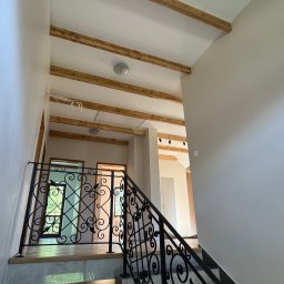 ArtBud - Wnętrze domu w trakcie wykańczania: schody z ozdobną balustradą, drewniane belki na suficie, widok na kolejne pomieszczenia. Neutralna kolorystyka ścian.
