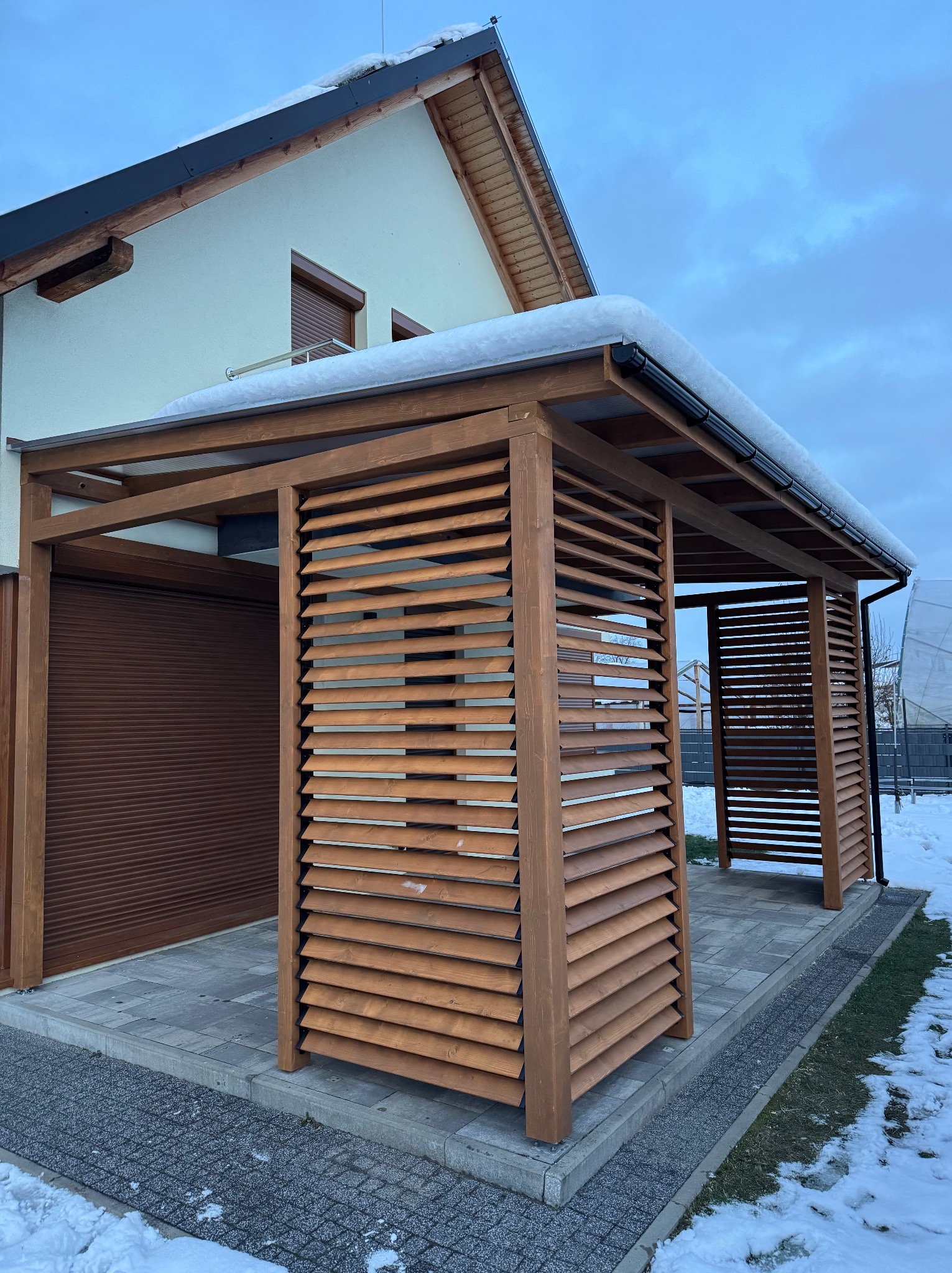Drewniana pergola z ruchomymi lamelami w kolorze złotego dębu, zadaszona poliwęglanem, na tle domu w zimowej scenerii. Widoczne detale konstrukcji i wykończenia.