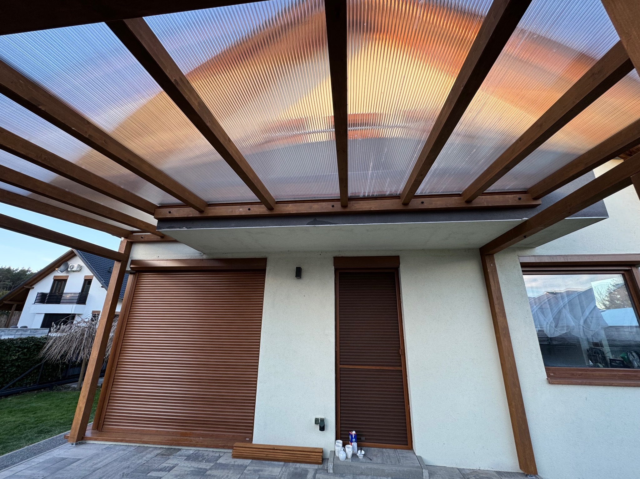 Drewniana pergola z przezroczystym zadaszeniem, widok od dołu, w tle budynek z roletami w Żarówce. Konstrukcja oparta o ścianę budynku.