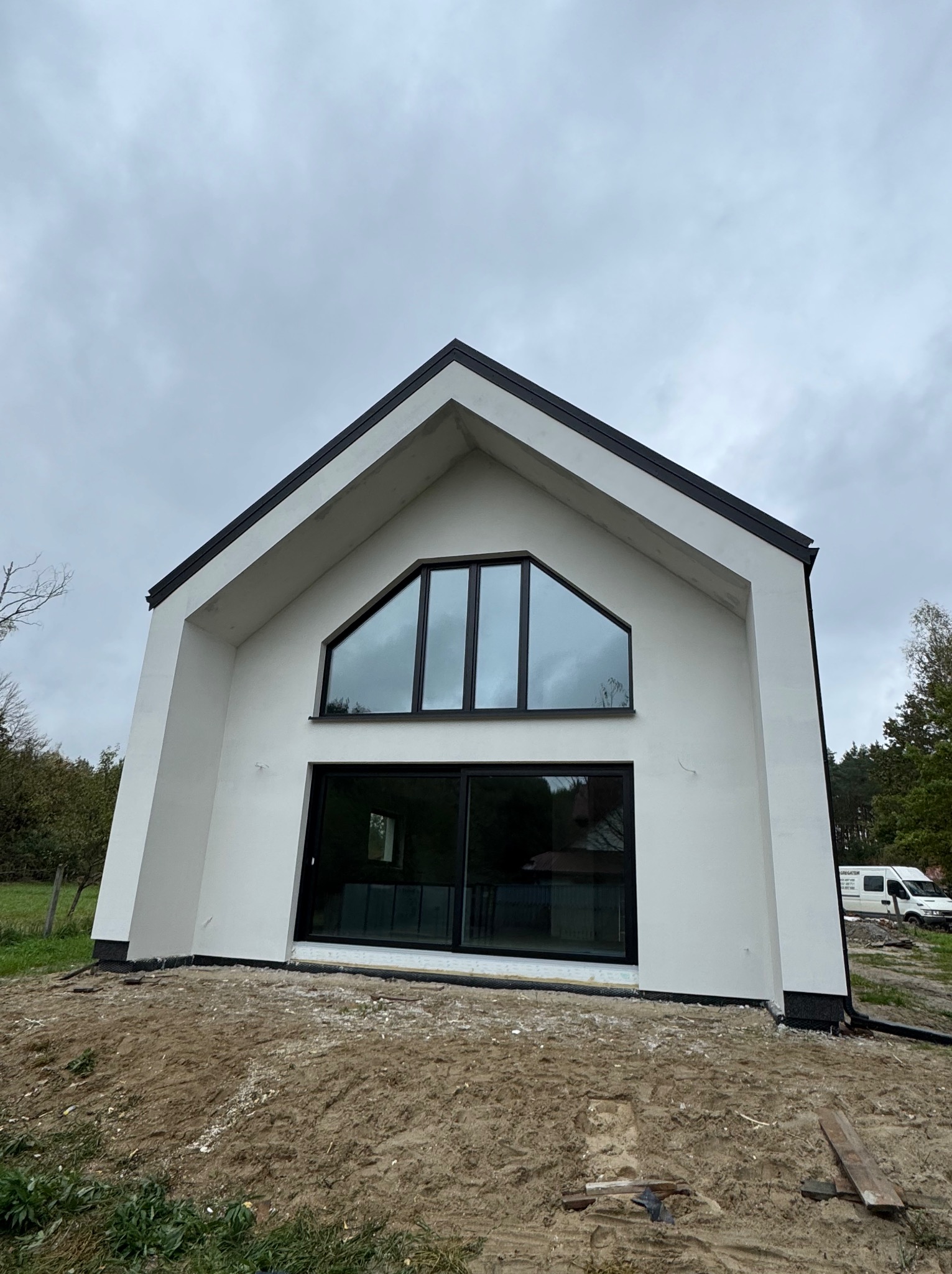 Nowoczesny dom z białymi ścianami i czarnymi ramami okiennymi. Charakterystyczne trójkątne okno na szczycie. Elewacja świeżo pomalowana, teren wokół w trakcie zagospodarowania.