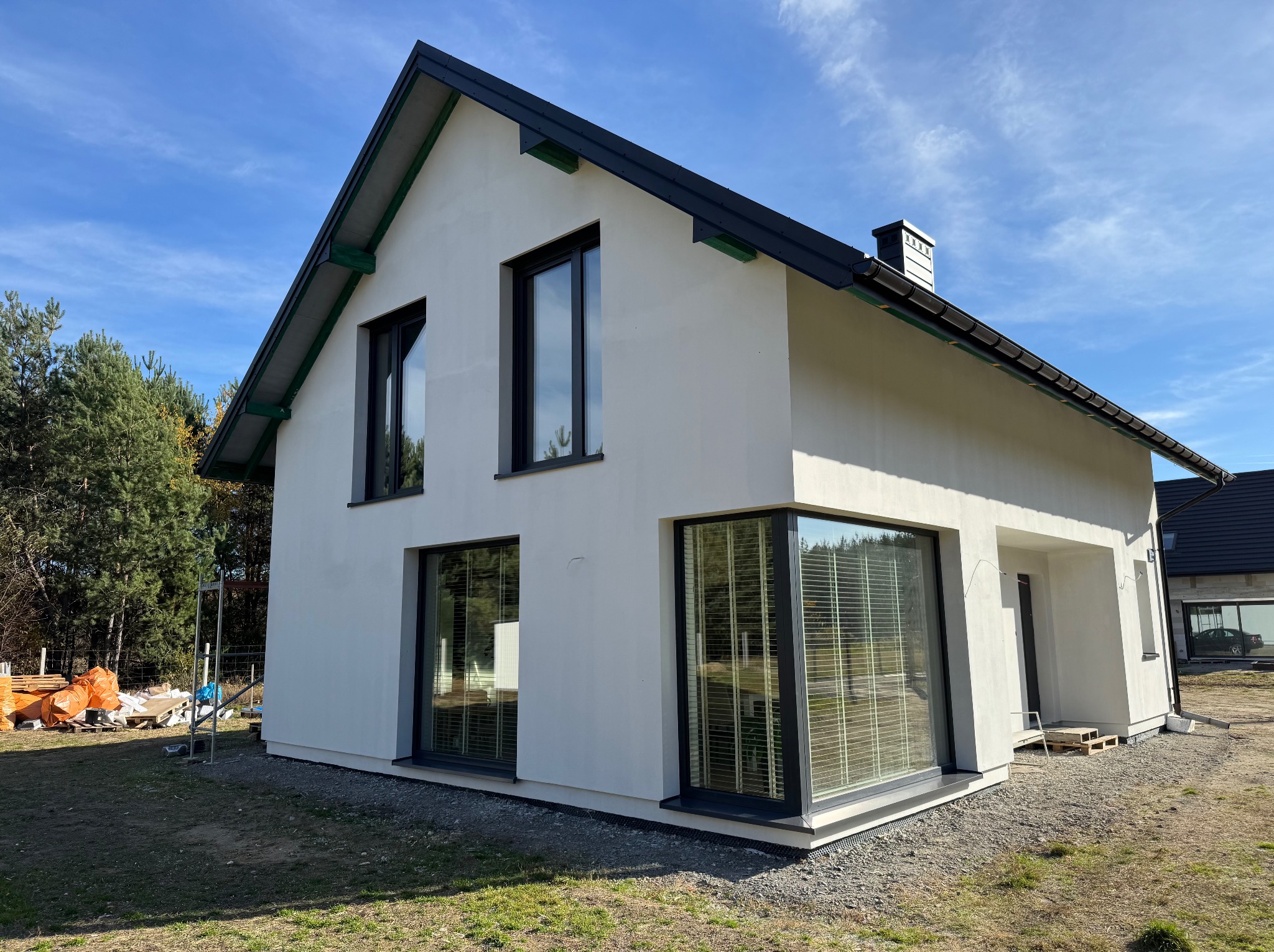 Nowoczesny, biały dom z dużymi oknami i czarnym dachem, otoczony zielenią i niebieskim niebem. Widoczne detale wykończenia elewacji i otoczenia.