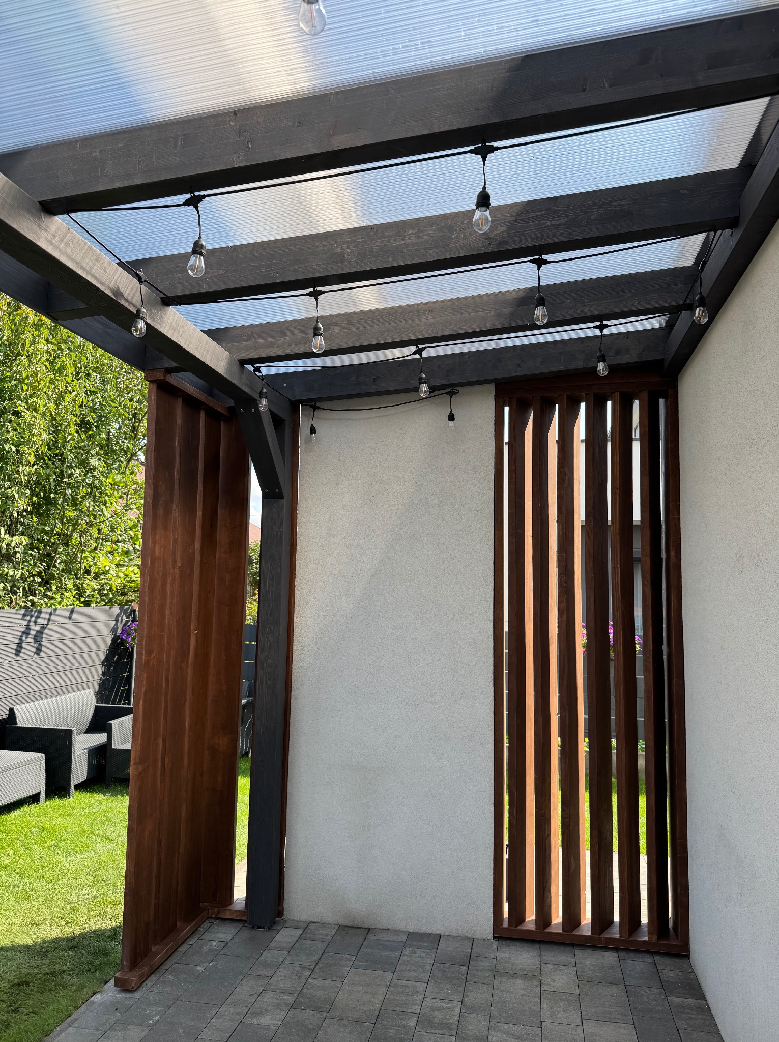 Nowoczesna pergola z ciemnego drewna z przezroczystym zadaszeniem i dekoracyjnym oświetleniem, przylegająca do białej ściany. Widoczne drewniane panele i szara kostka brukowa.