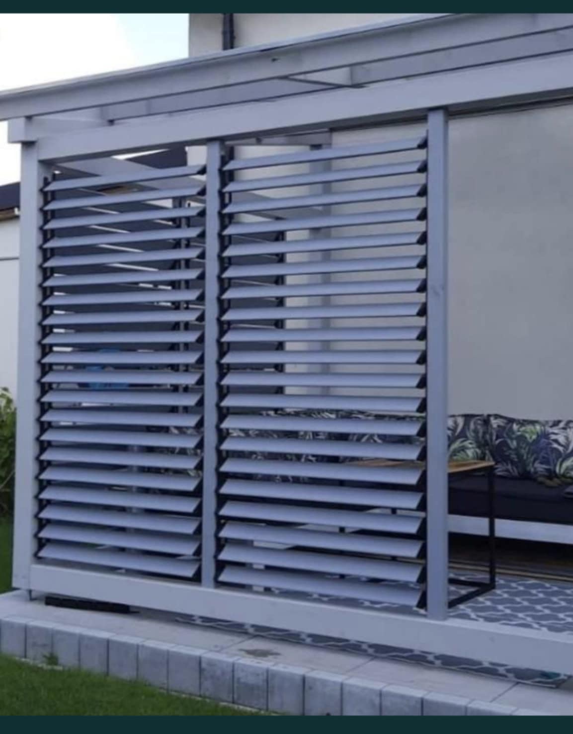 Nowoczesna, szara pergola metalowa z regulowanymi lamelami, przylegająca do budynku. Widoczny fragment sofy z poduszkami w tle.