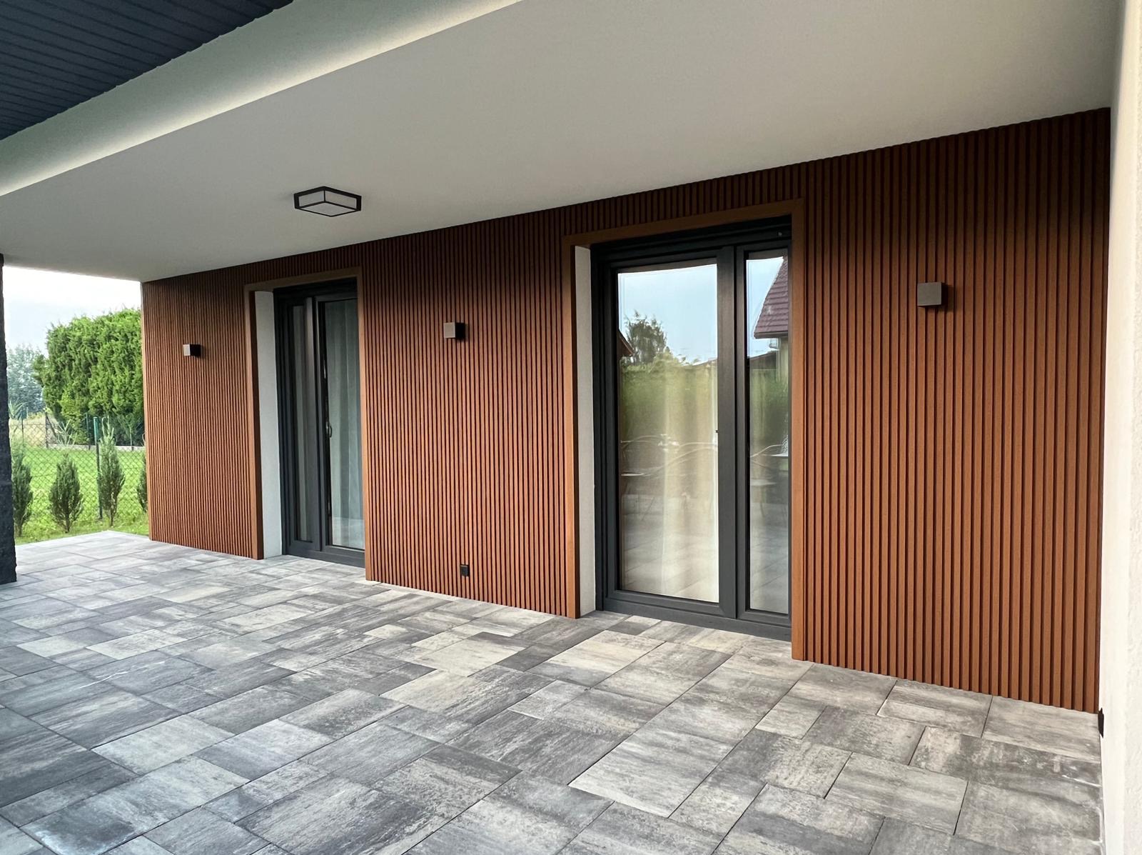Nowoczesny taras z szarej kostki brukowej i elewacją z paneli imitujących drewno. Dwa przesuwne okna z czarnymi ramami. Oświetlenie zewnętrzne.