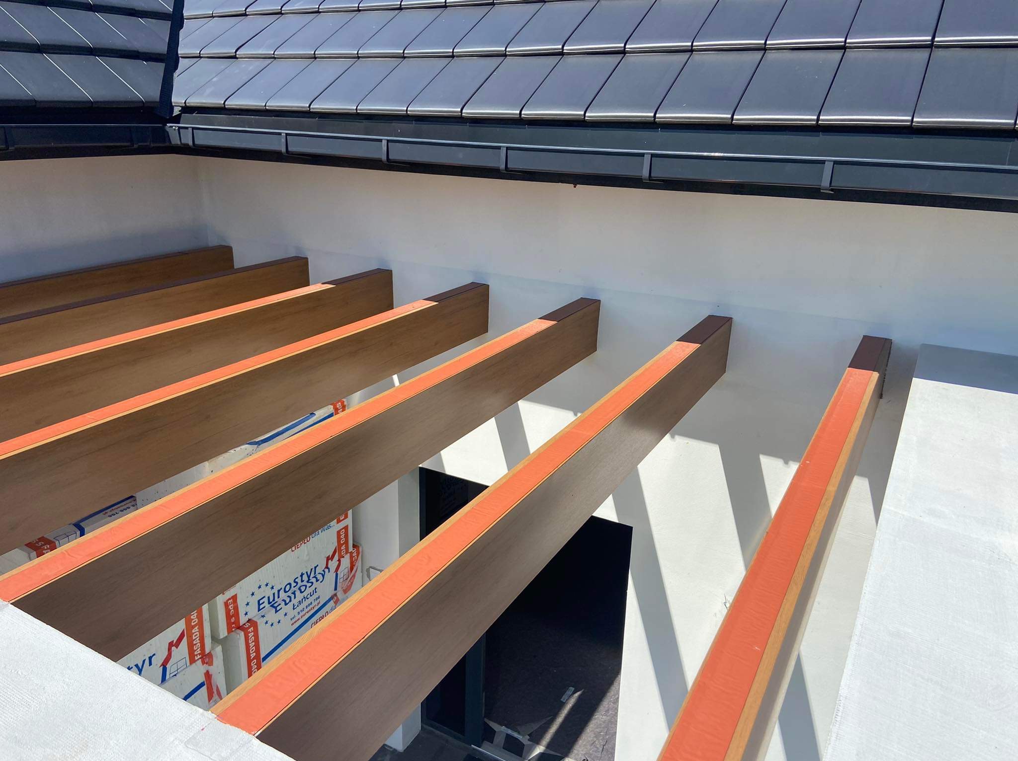 Drewniana pergola z pomarańczowymi akcentami, widok z dołu na tle białej ściany i fragmentu dachu z czarnej dachówki. Widoczne ocieplenie między belkami.