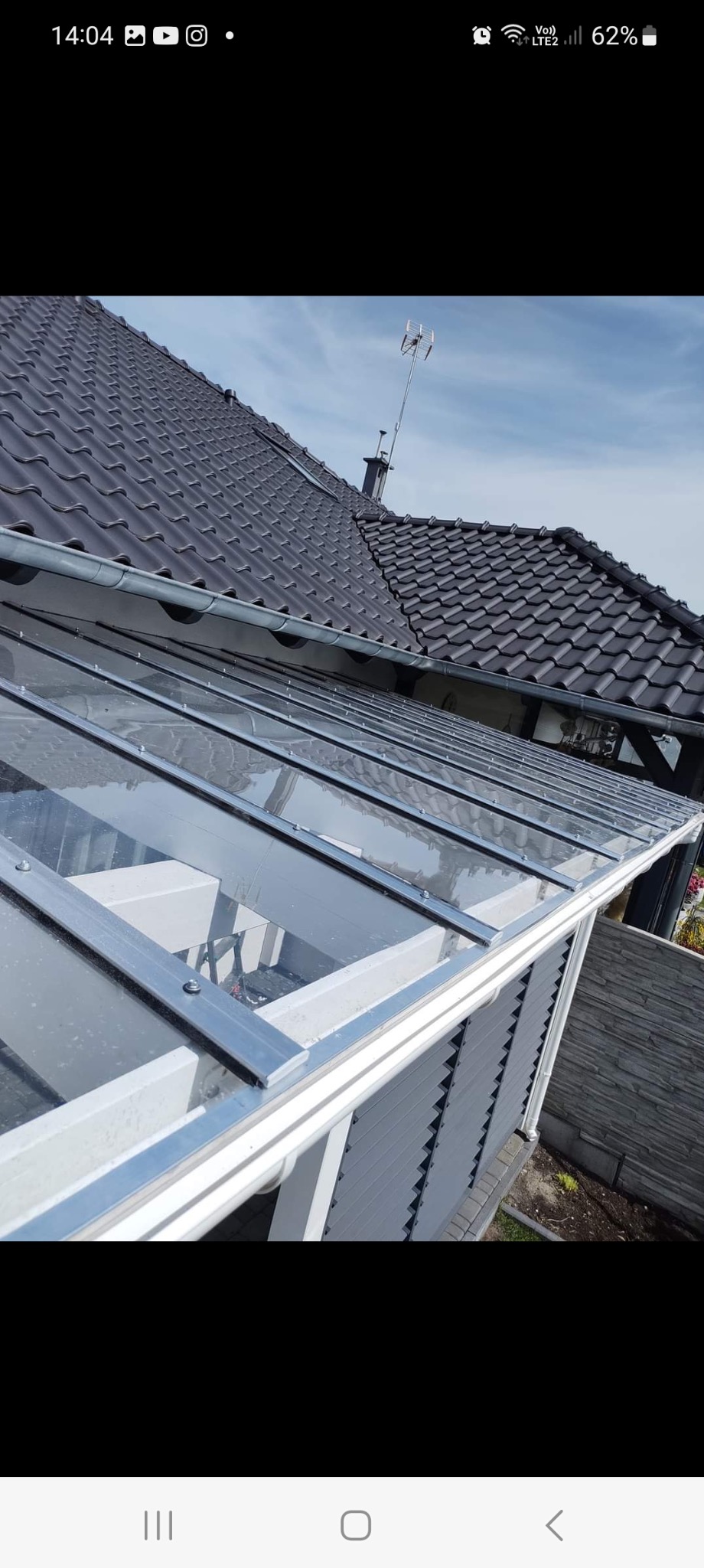 Szklana zabudowa tarasu z metalową konstrukcją, widok z góry, z fragmentem dachu domu w tle. Nowoczesne rozwiązanie zadaszenia.