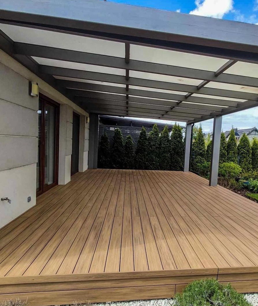 Nowoczesny taras z drewnianą podłogą i metalową pergolą, otoczony zielenią. Widok na dom z dużymi oknami i starannie utrzymany ogród z tujami.