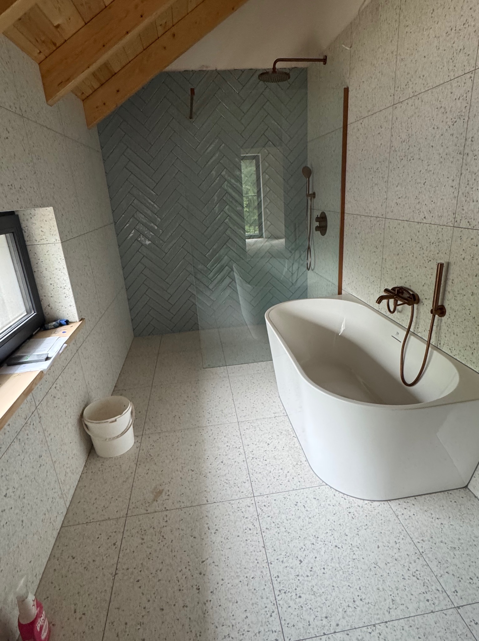 Nowoczesna łazienka na poddaszu z wanną wolnostojącą, szklaną kabiną prysznicową i mozaiką w jodełkę. Widoczny kubeł i butelka chemii. Ukończone wykończenie.