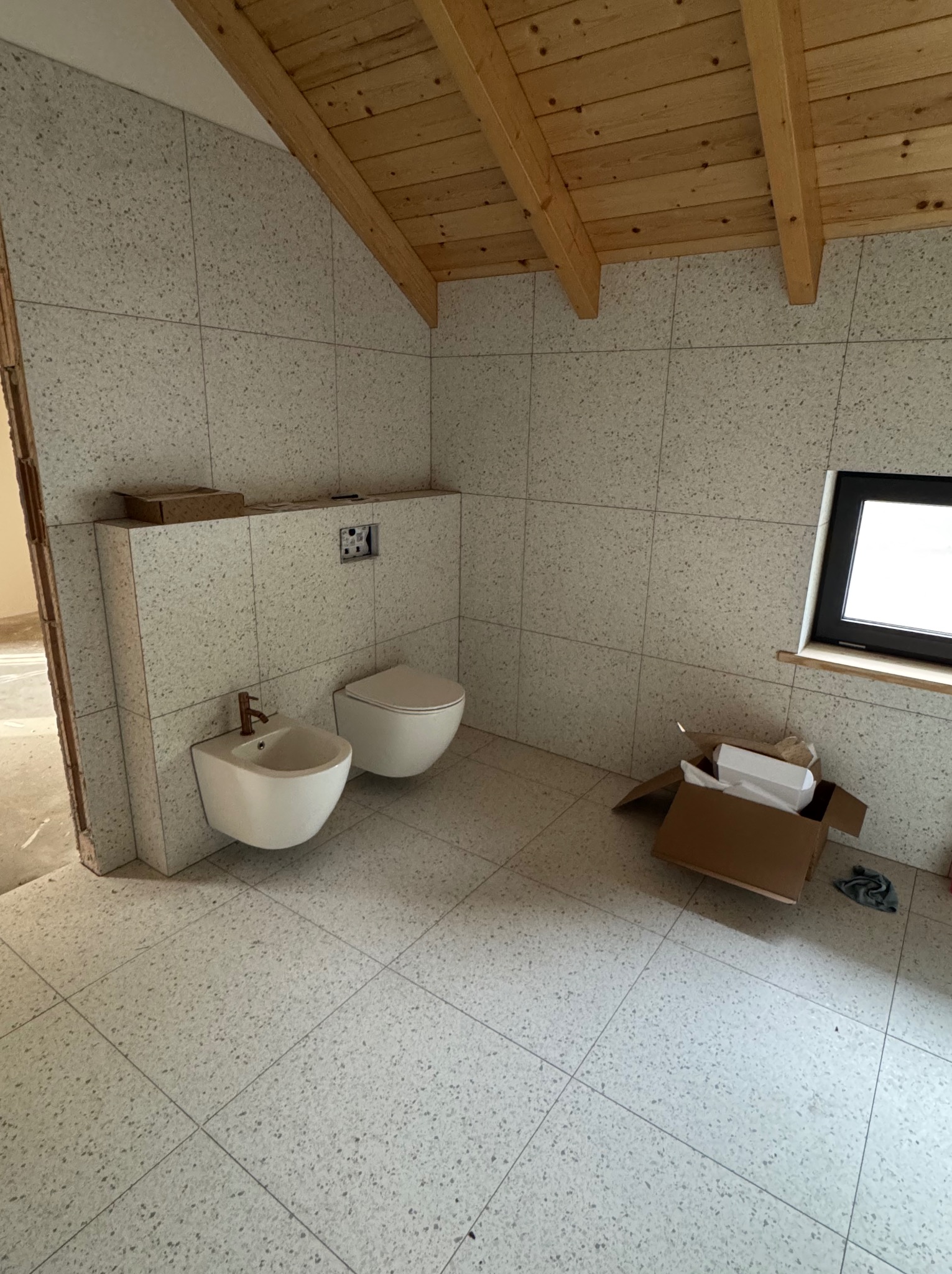 Wykończona łazienka na poddaszu z beżowymi płytkami, widoczne białe sanitariaty, drewniana więźba dachowa i okno. Częściowo widoczny karton po armaturze.