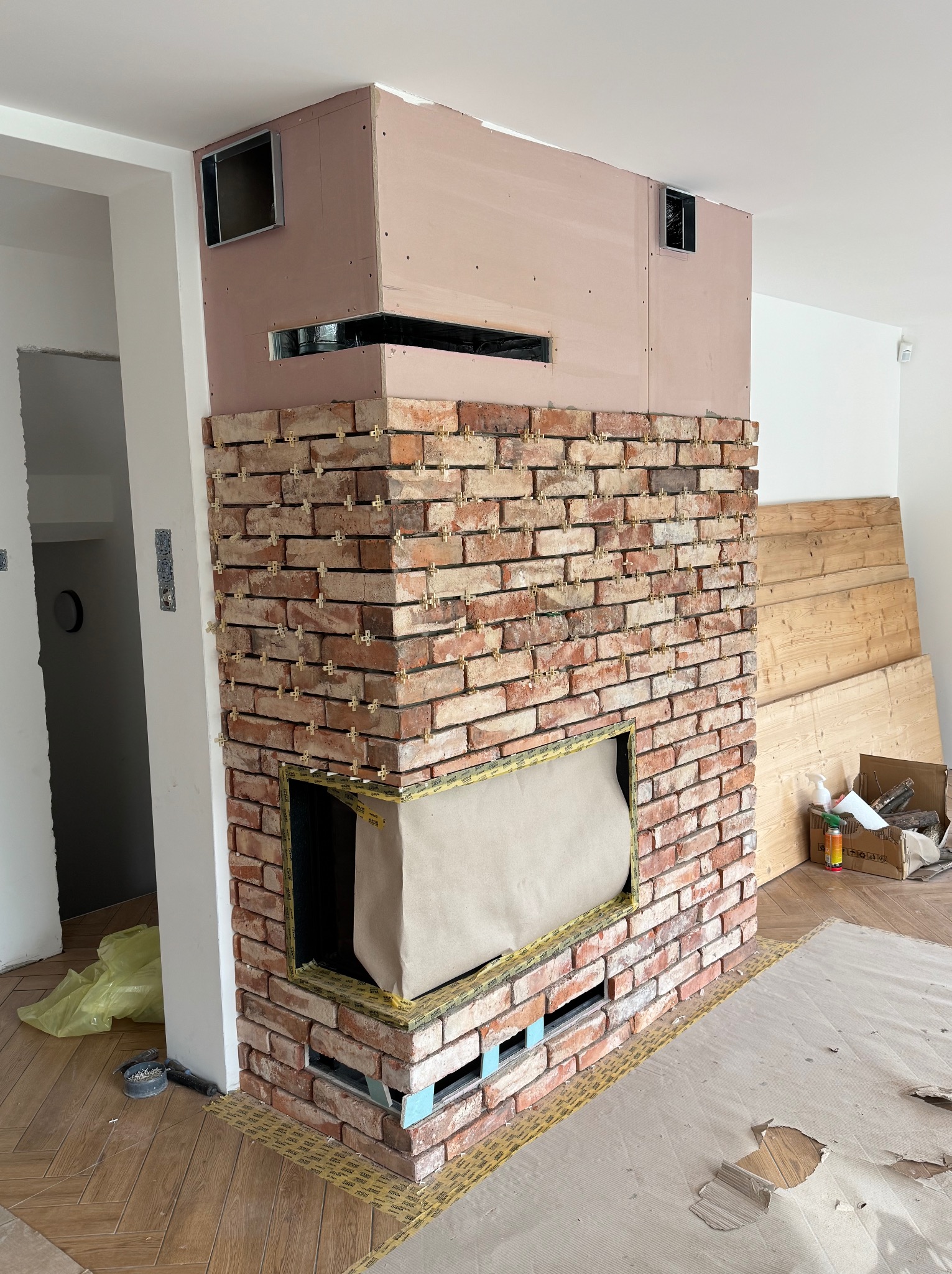 Wnętrze w trakcie prac: obudowa kominka z cegły klinkierowej, widoczne krzyżyki dystansowe, górna część obudowy w surowym stanie, zabezpieczona folią podłoga.