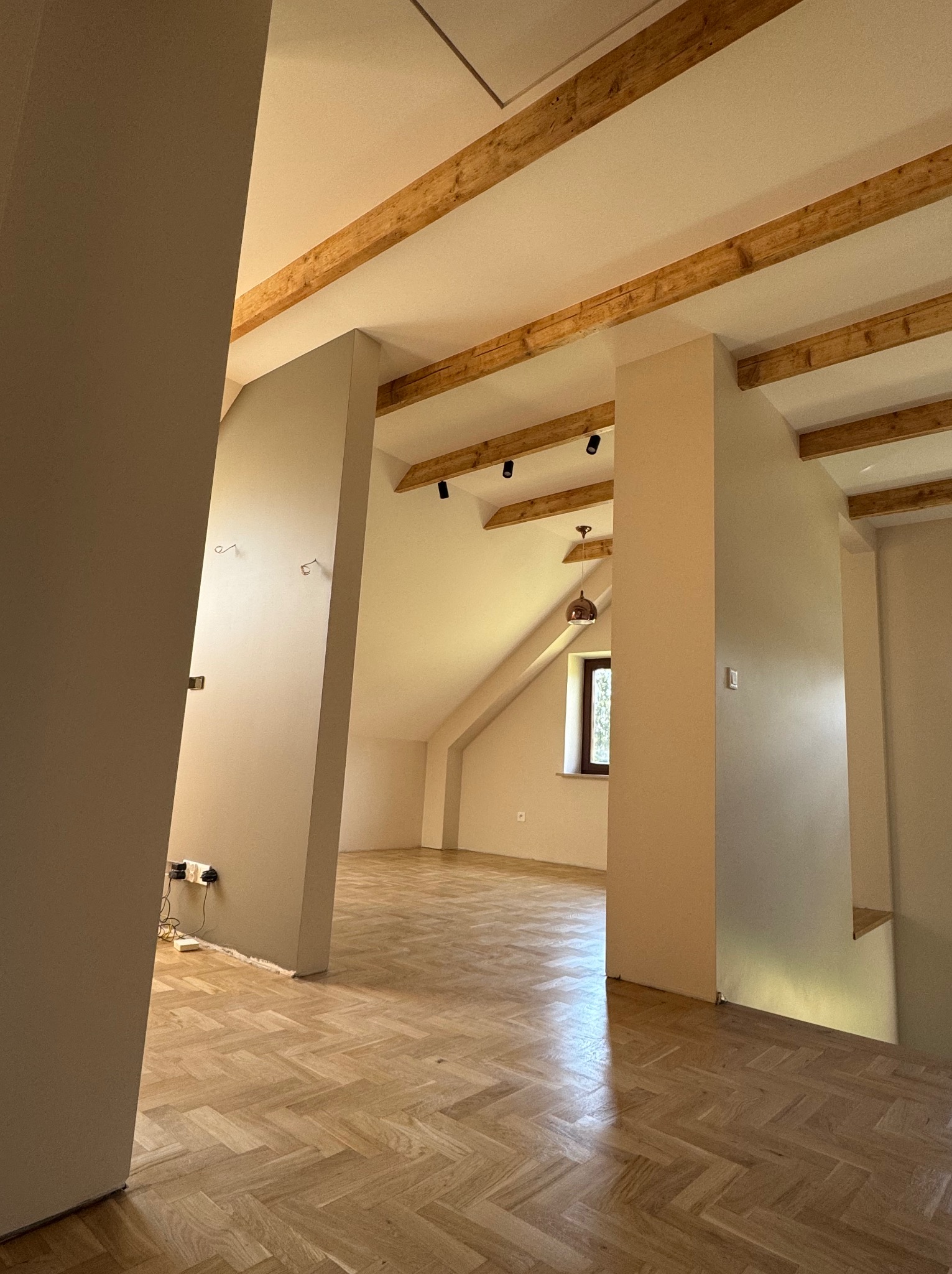 Jasne, przestronne poddasze po remoncie z drewnianą podłogą w jodełkę, beżowymi ścianami i drewnianymi belkami stropowymi. Widoczne gniazdka elektryczne i lampy.