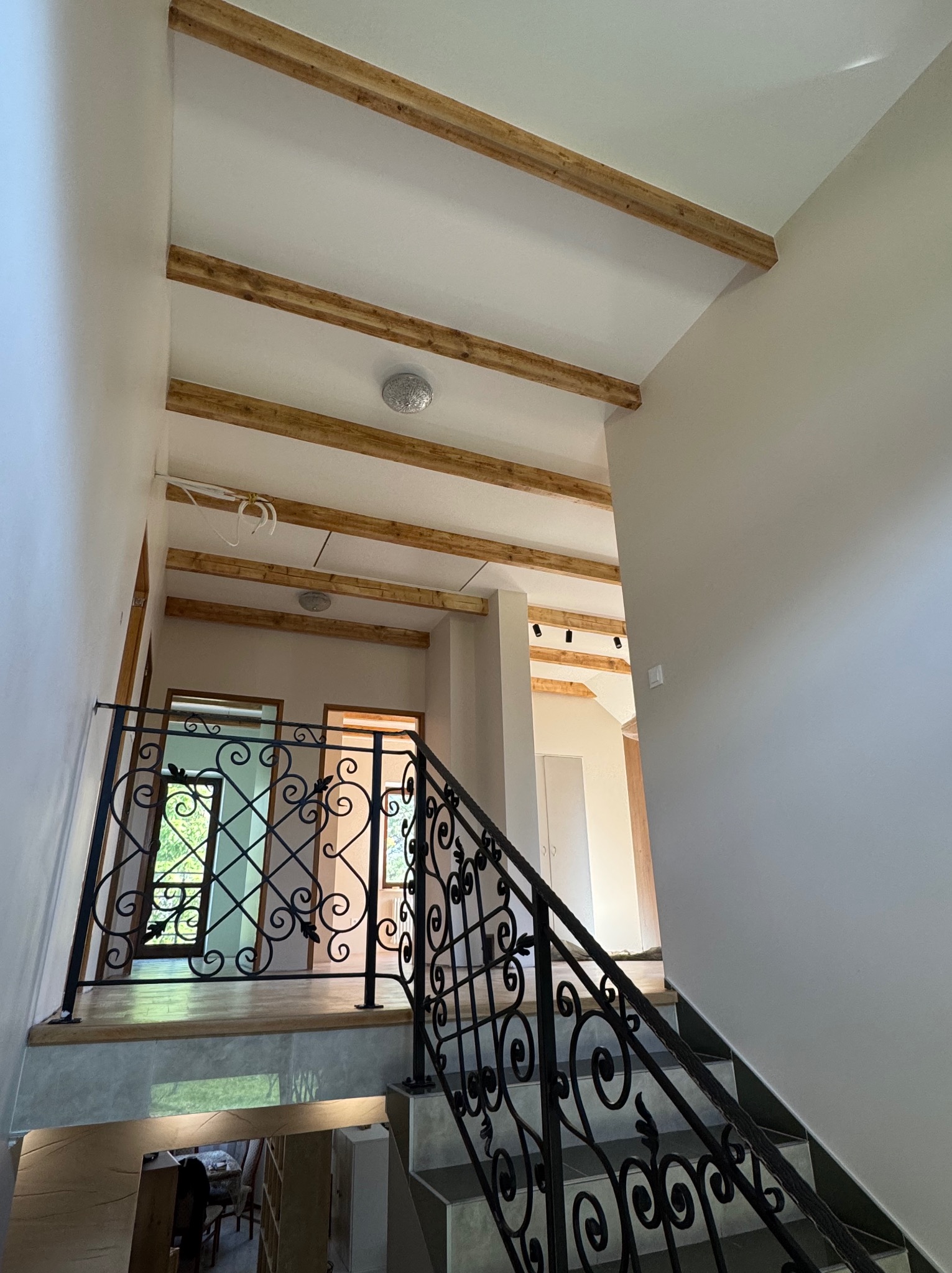 Wnętrze domu w trakcie wykańczania: schody z ozdobną balustradą, drewniane belki na suficie, widok na kolejne pomieszczenia. Neutralna kolorystyka ścian.