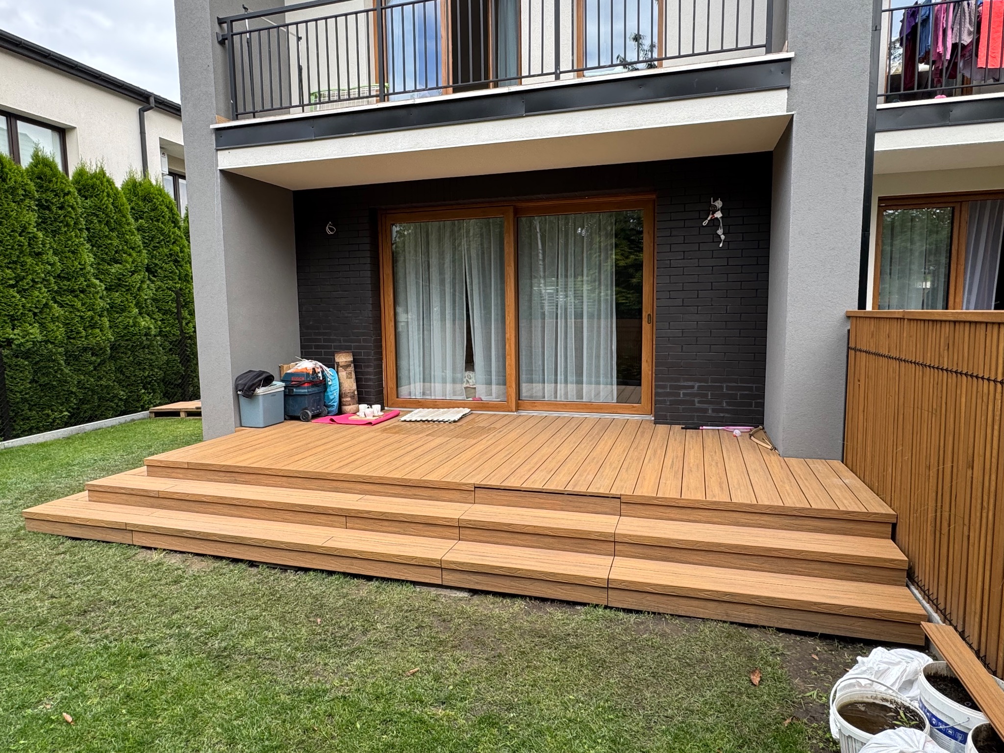 Nowoczesny taras kompozytowy z trzema stopniami, prowadzący do drzwi balkonowych. Widoczne elementy wykończeniowe i fragment ogrodu z tujami.