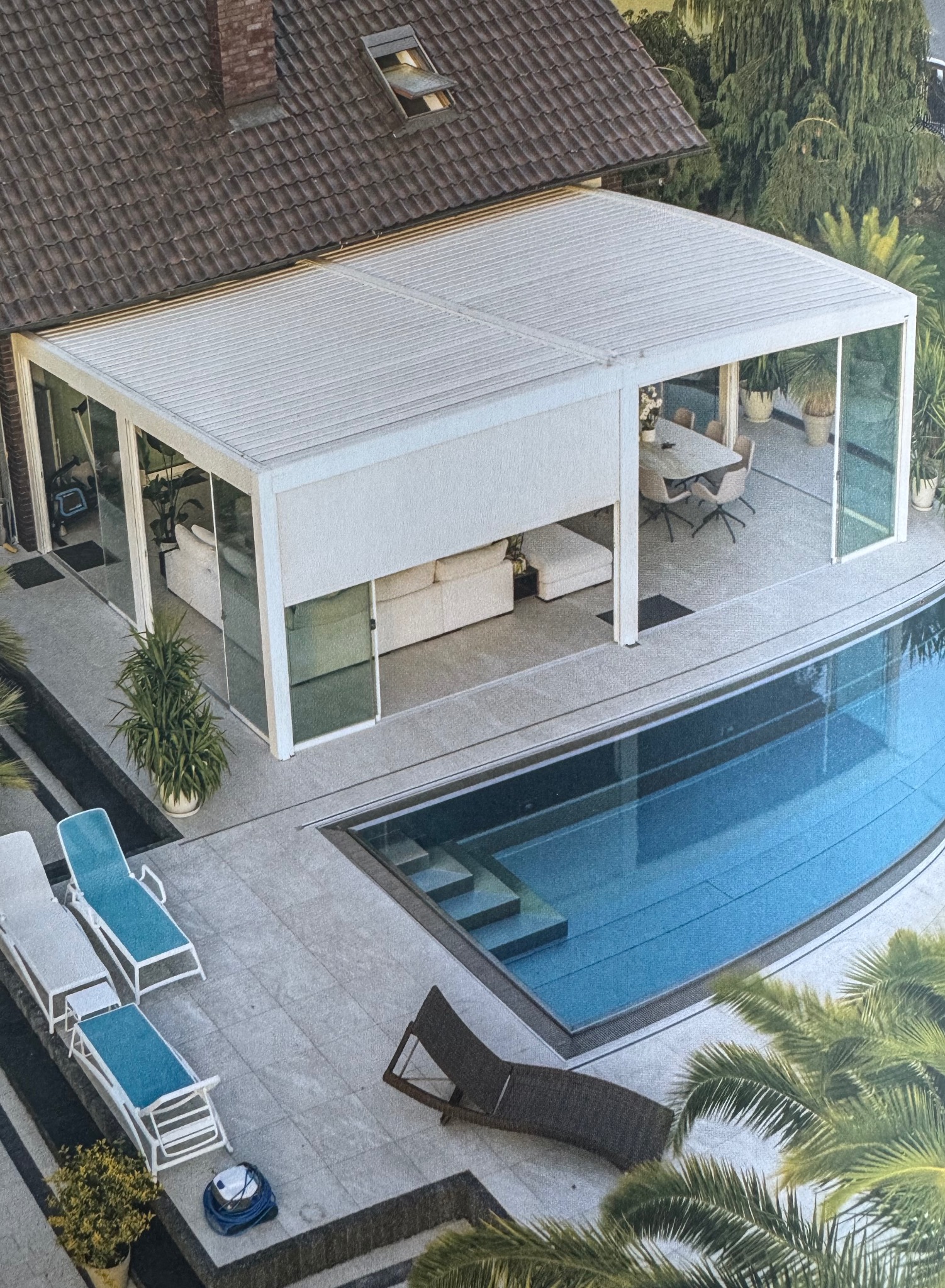 Nowoczesna, przeszklona pergola z białymi ramami, przylegająca do domu z basenem. Widoczne meble ogrodowe i leżaki. Ujęcie z góry, podkreślające przestronność i luksusowy charakter.