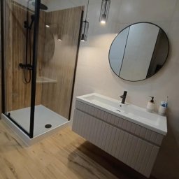 REMONT4YOU Bartłomiej Nikrant - Nowoczesna łazienka z kabiną prysznicową typu walk-in z czarną ramą, wiszącą szafką z umywalką i okrągłym lustrem. Drewnopodobne płytki na podłodze i ścianie kabiny.