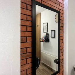 REMONT4YOU Bartłomiej Nikrant - Ściana z cegły klinkierowej z czarnym lustrem, odbijającym fragment wnętrza z lamelami, półką i grzejnikiem. Oświetlenie punktowe na ścianie.