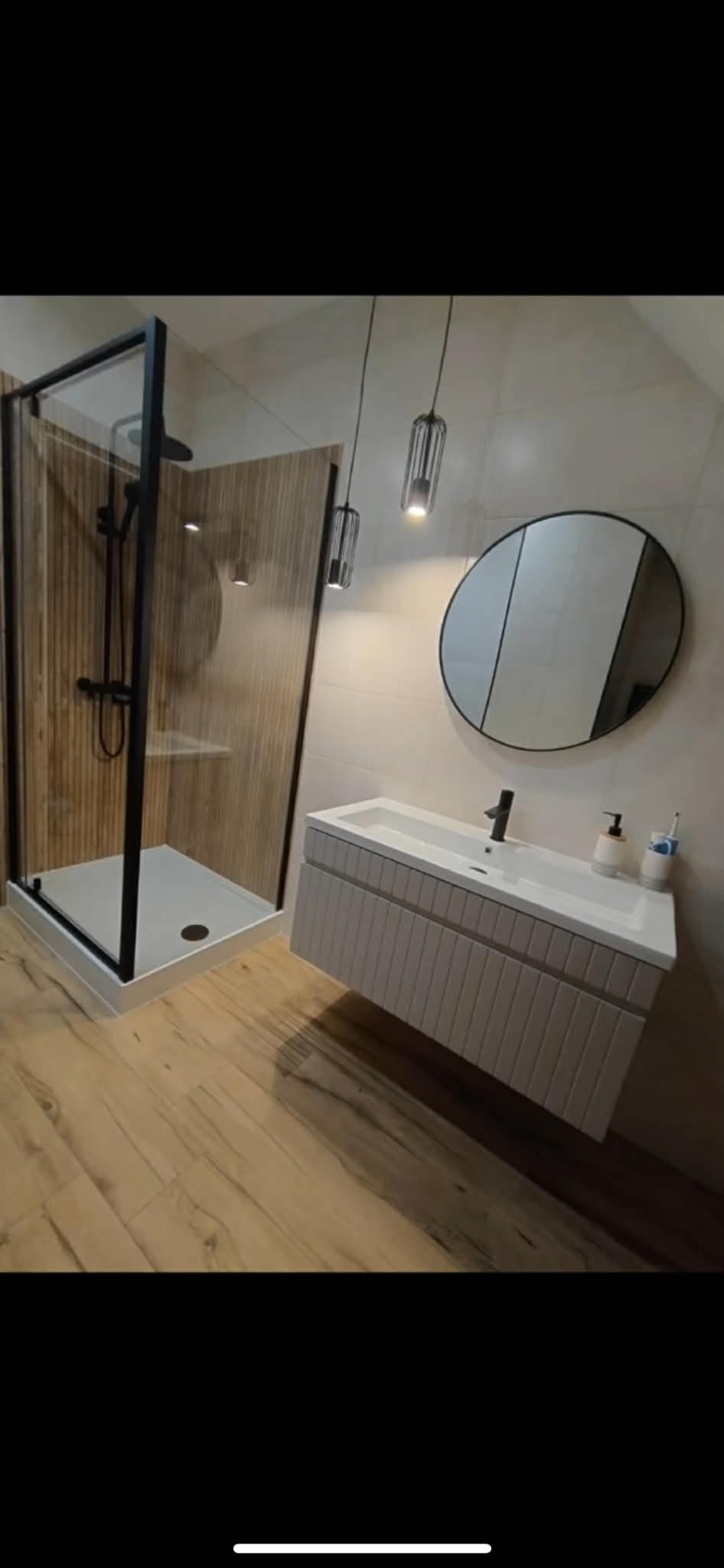 Nowoczesna łazienka z kabiną prysznicową typu walk-in z czarną ramą, wiszącą szafką z umywalką i okrągłym lustrem. Drewnopodobne płytki na podłodze i ścianie kabiny.